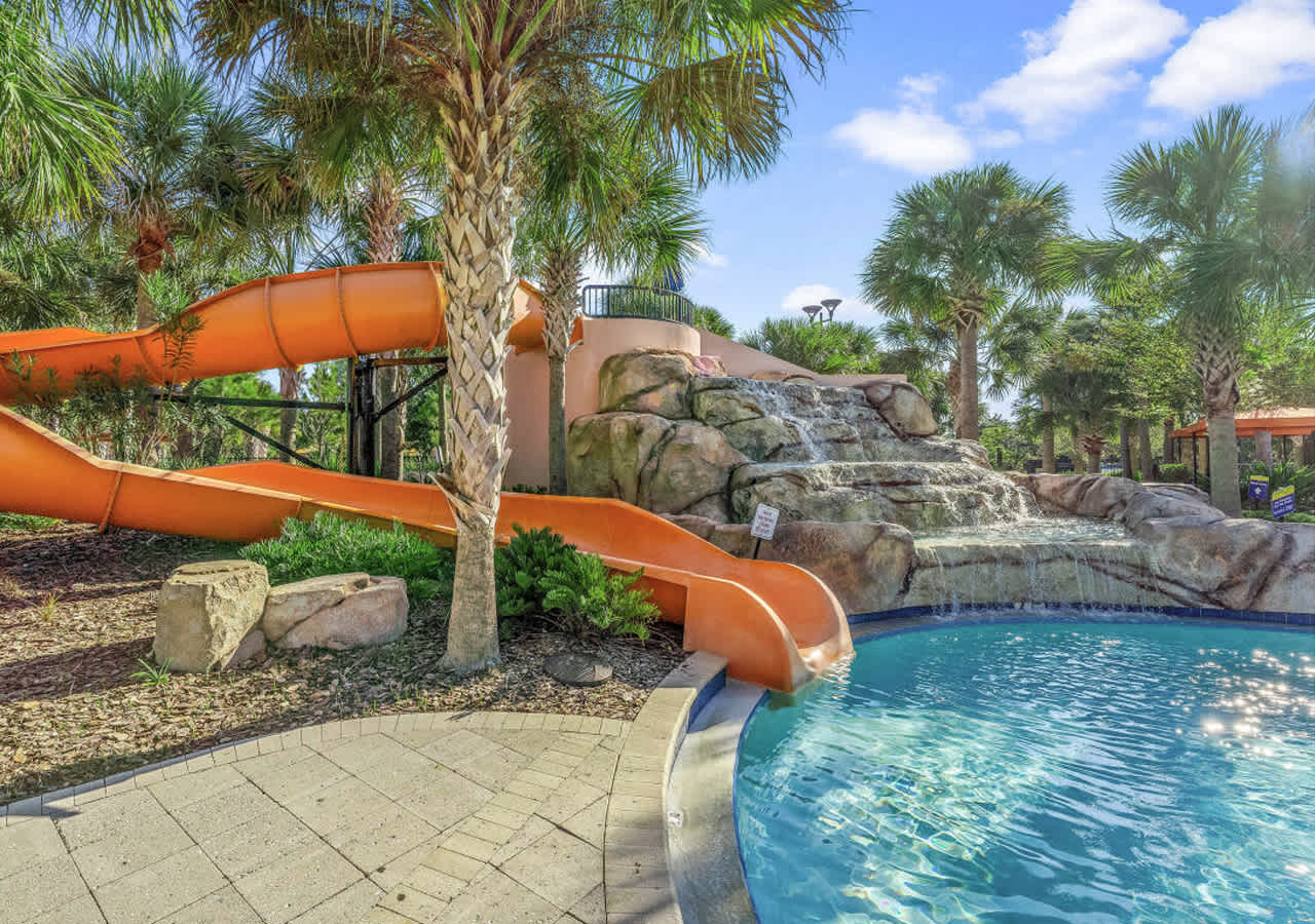 Bright orange water slides curve down into a clear blue pool, complemented by a natural-looking rock waterfall feature and surrounded by lush palm trees, creating an exciting water play area.