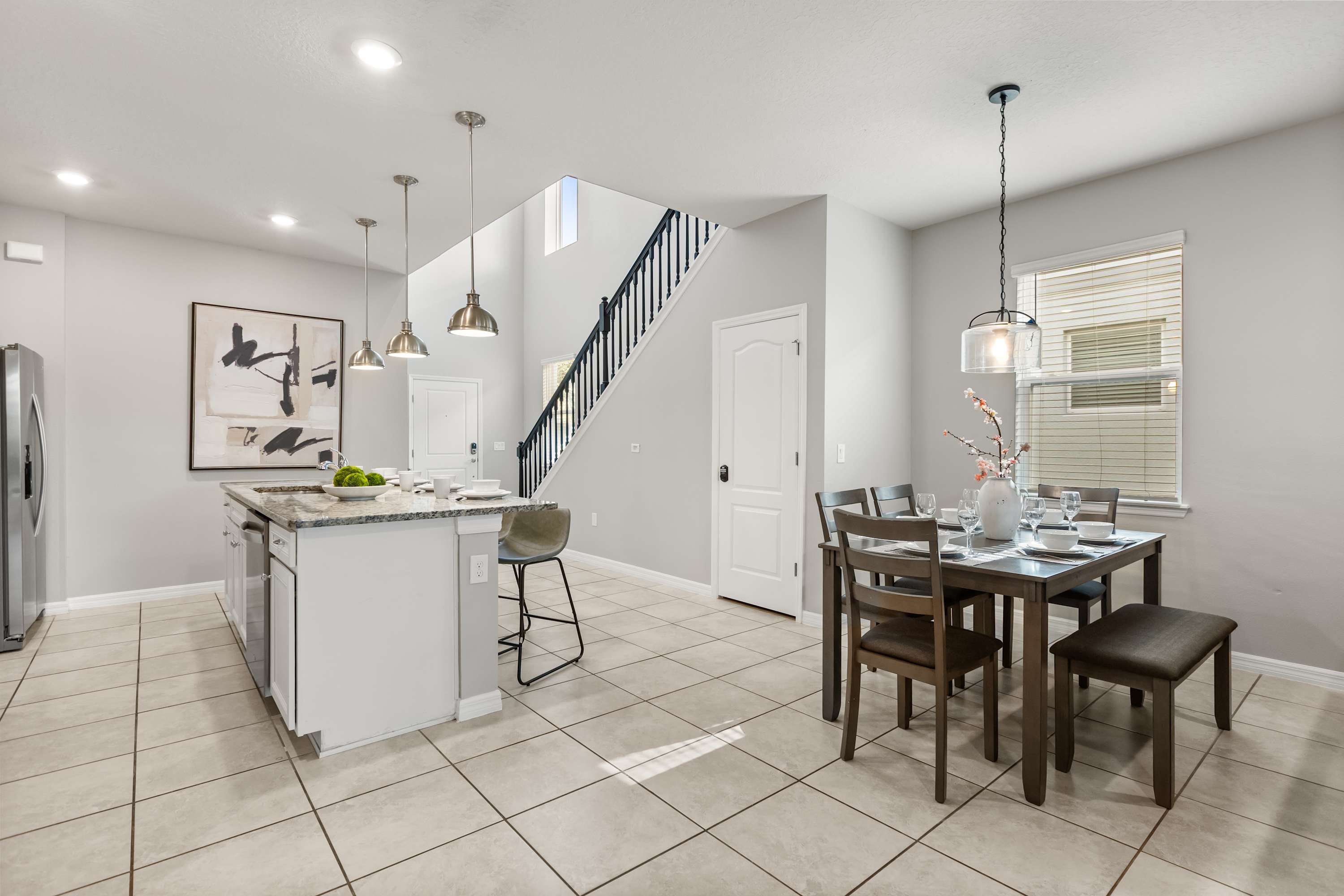 Open-concept dining area with a dark wood dining table and chairs, set on light tile flooring. The kitchen island with bar seating is visible to the left, and a staircase leads upstairs in the background.