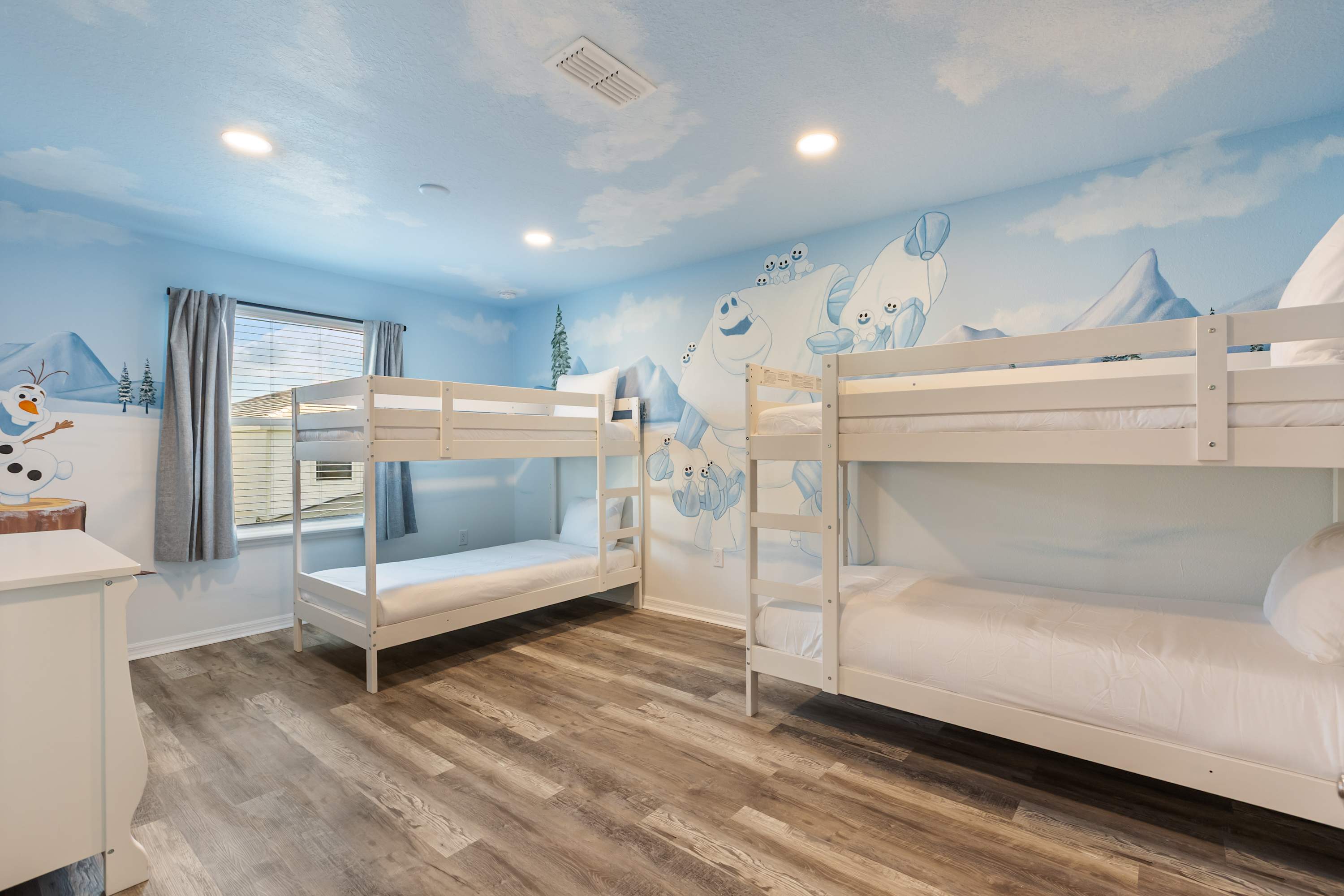A wide shot of the "Frozen" themed bedroom, featuring two white bunk beds with white bedding and a playful mural of snowmen on the far wall. The room has wood-look flooring and a window with blinds.