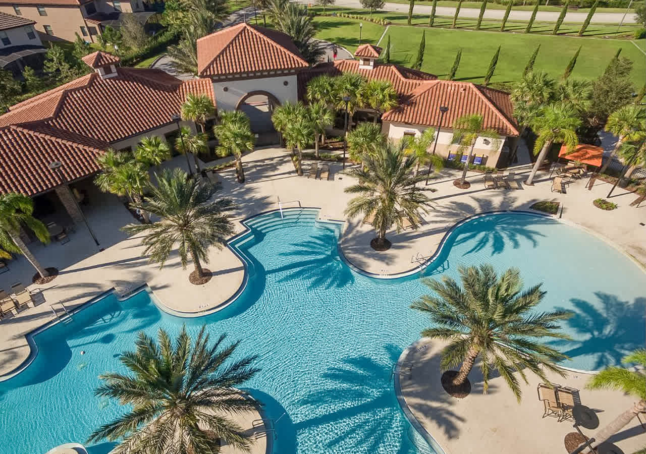 An aerial view of a large, irregularly shaped community swimming pool with bright blue water, surrounded by palm trees and lounge chairs. In the background, a clubhouse with terracotta roofs and lush green fields are visible under a sunny sky.