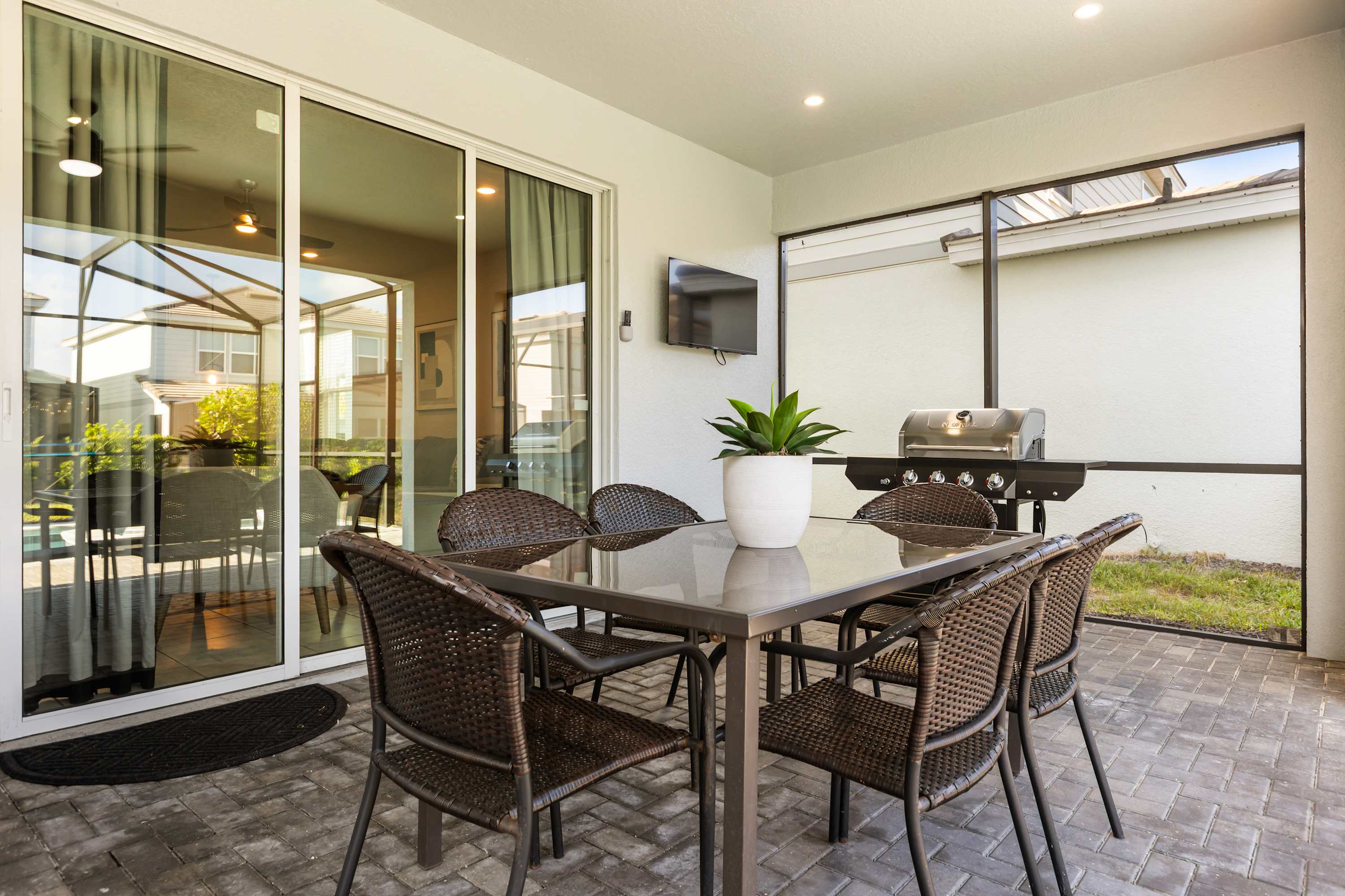 A covered outdoor patio features a dark square dining table with six wicker chairs and a BBQ grill, all set on brick pavers. A wall-mounted TV is visible, and the area is enclosed by screening.