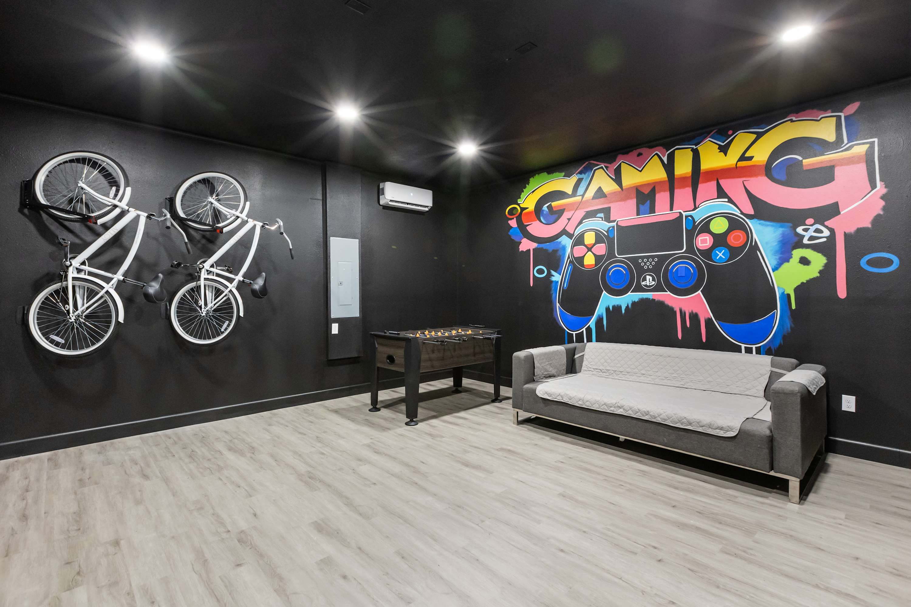 A black-walled game room features a vibrant "GAMING" mural, a foosball table, and a gray sofa. Two white bicycles are mounted decoratively on the wall, and a mini-split AC unit is visible.