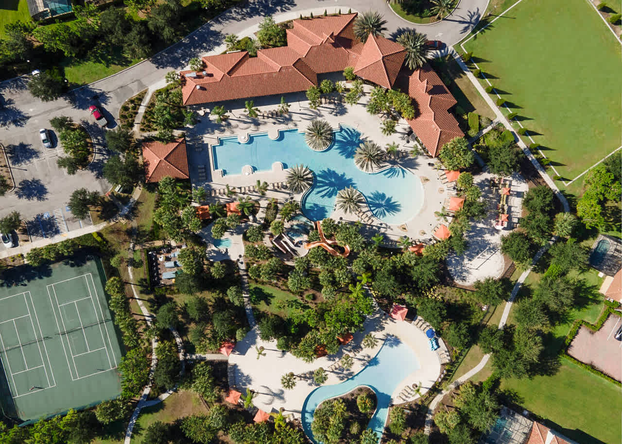 A high aerial perspective captures the extensive resort amenity area, highlighting a large pool complex with a lazy river, water slides, and a tennis court, all nestled within a landscaped community with residential homes in the distance.