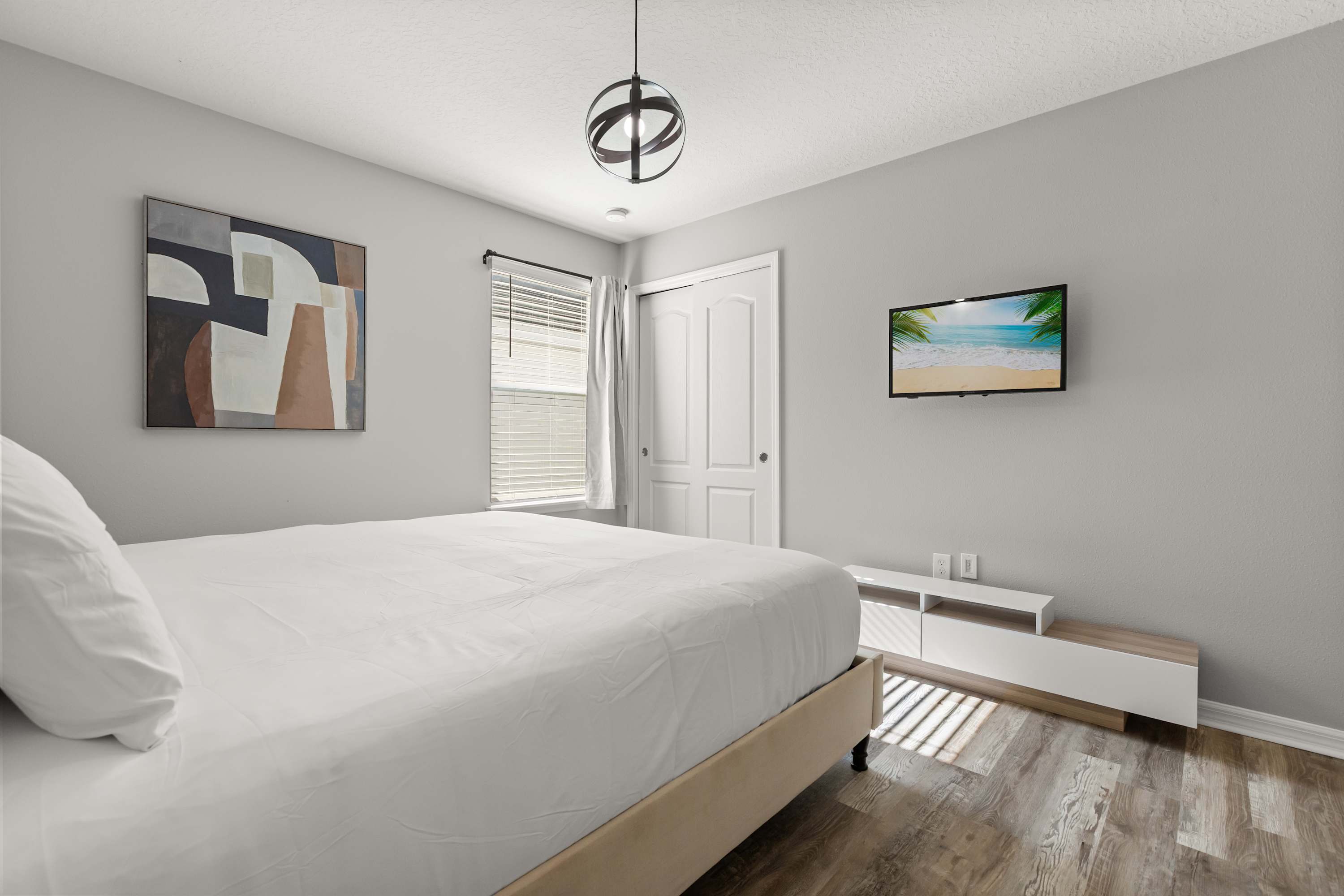 A bright bedroom with a large bed covered in white bedding, light gray walls, and wood-look flooring. A wall-mounted TV is positioned above a low white media console, and a window with blinds and curtains allows natural light.