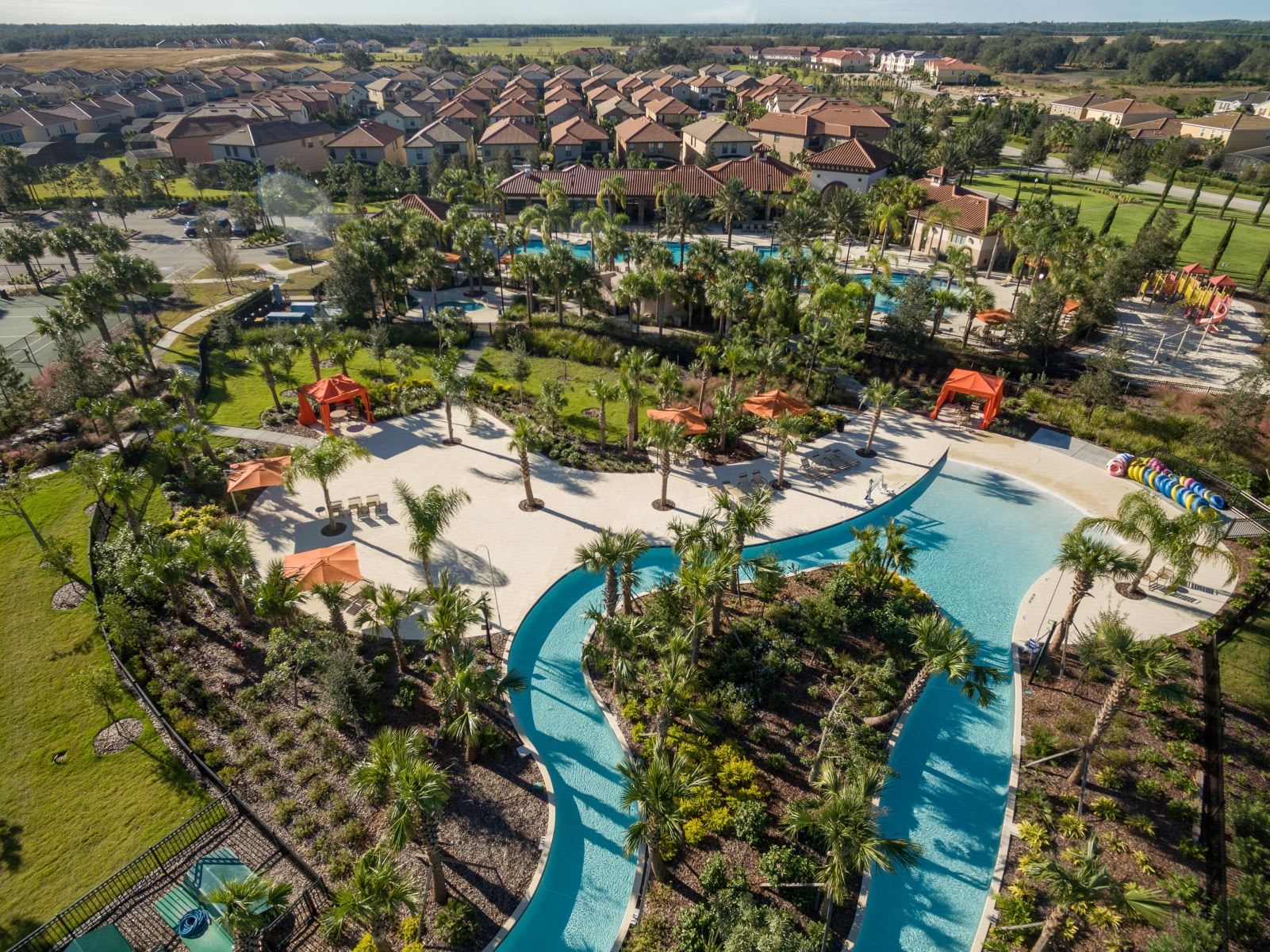 An aerial shot focuses on the winding lazy river amenity, bordered by lush landscaping, palm trees, and orange sun umbrellas, with a residential community of houses visible in the background under a bright sky.