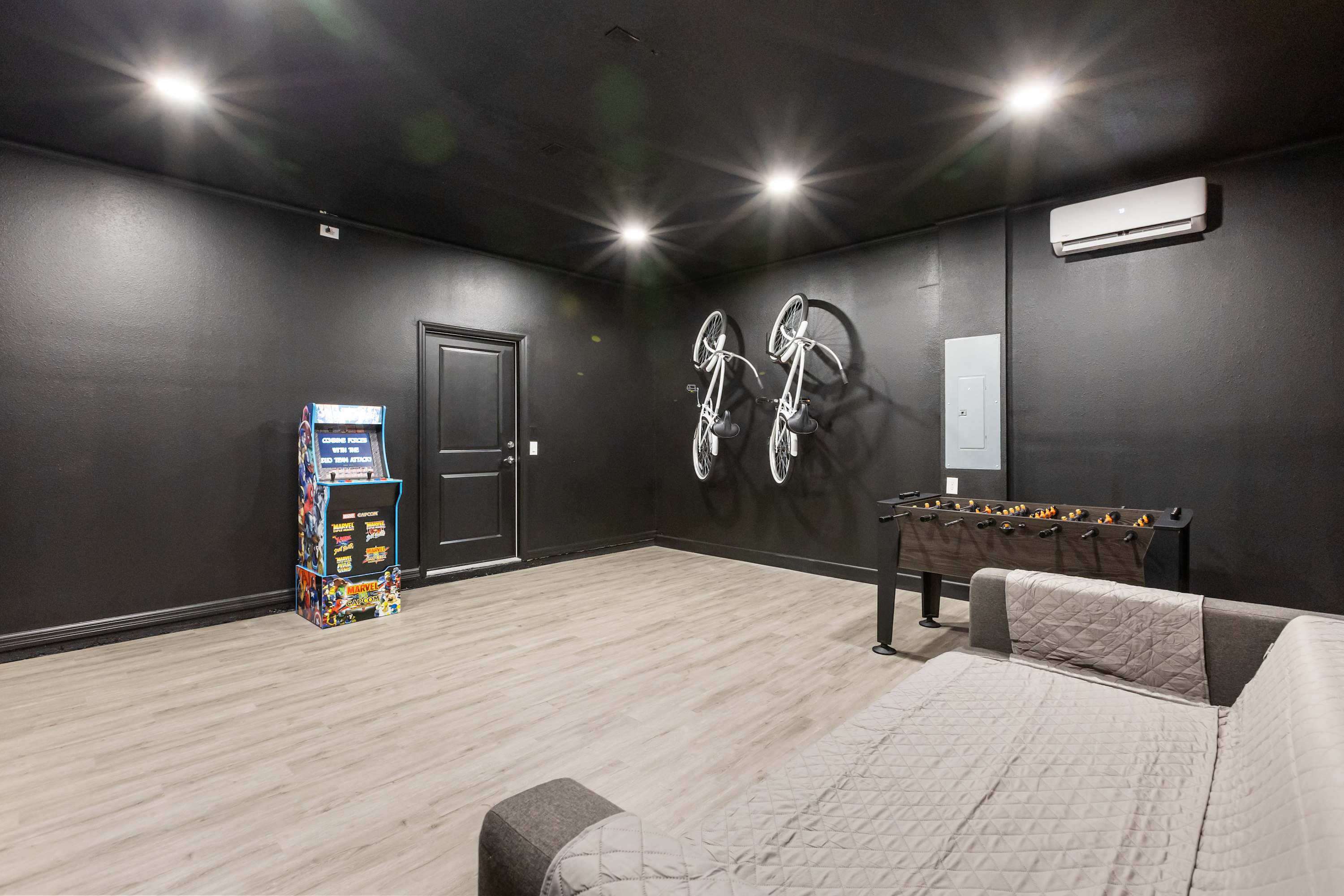 A corner of the black-walled game room features a classic arcade machine and a dark door, with two white bicycles mounted on the wall and a foosball table visible in the background. The room has light wood-look flooring.