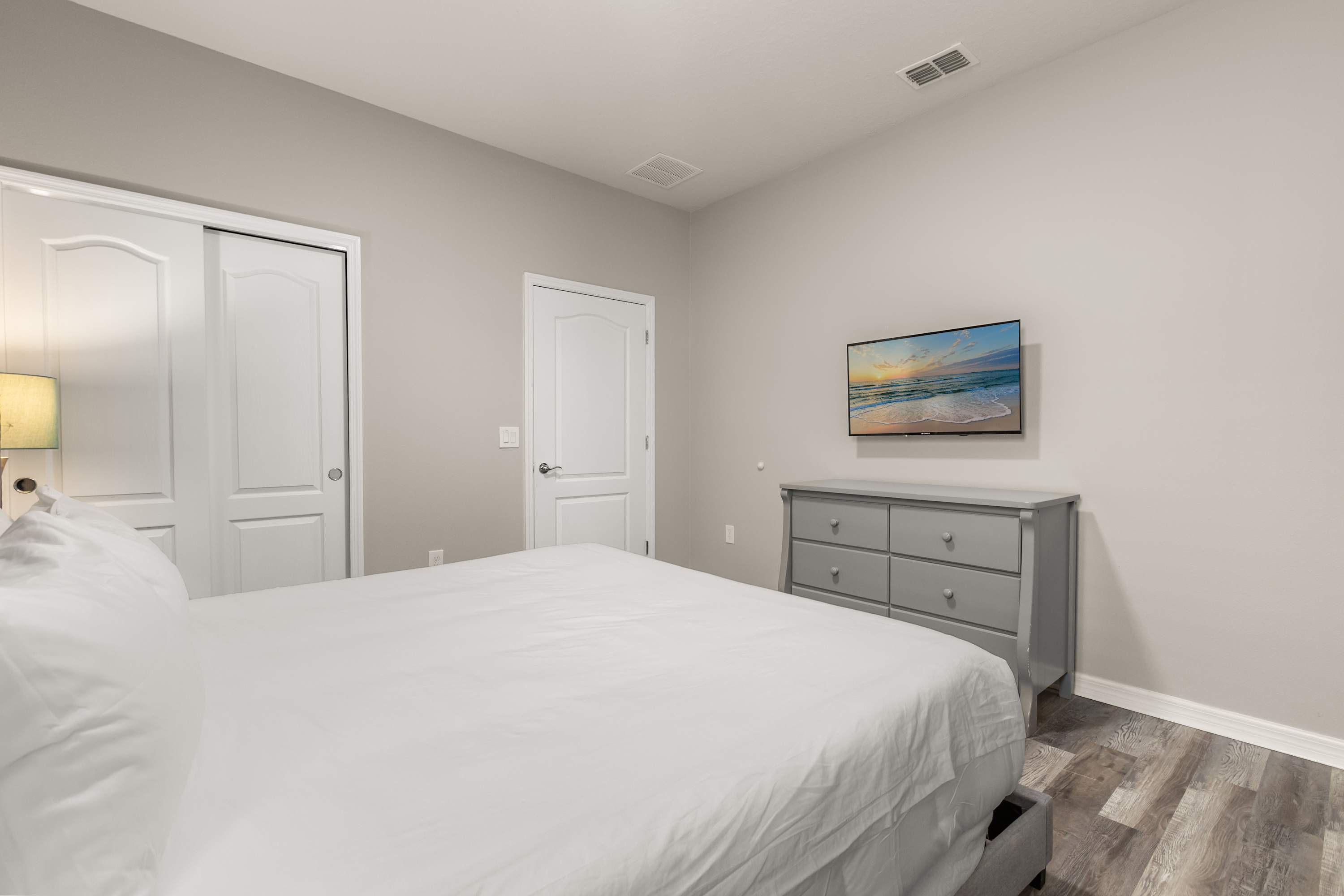 View of a bedroom with a gray upholstered queen bed, light gray walls, and wood-look laminate flooring. A wall-mounted TV is positioned above a gray dresser, and white closet doors are visible to the left.