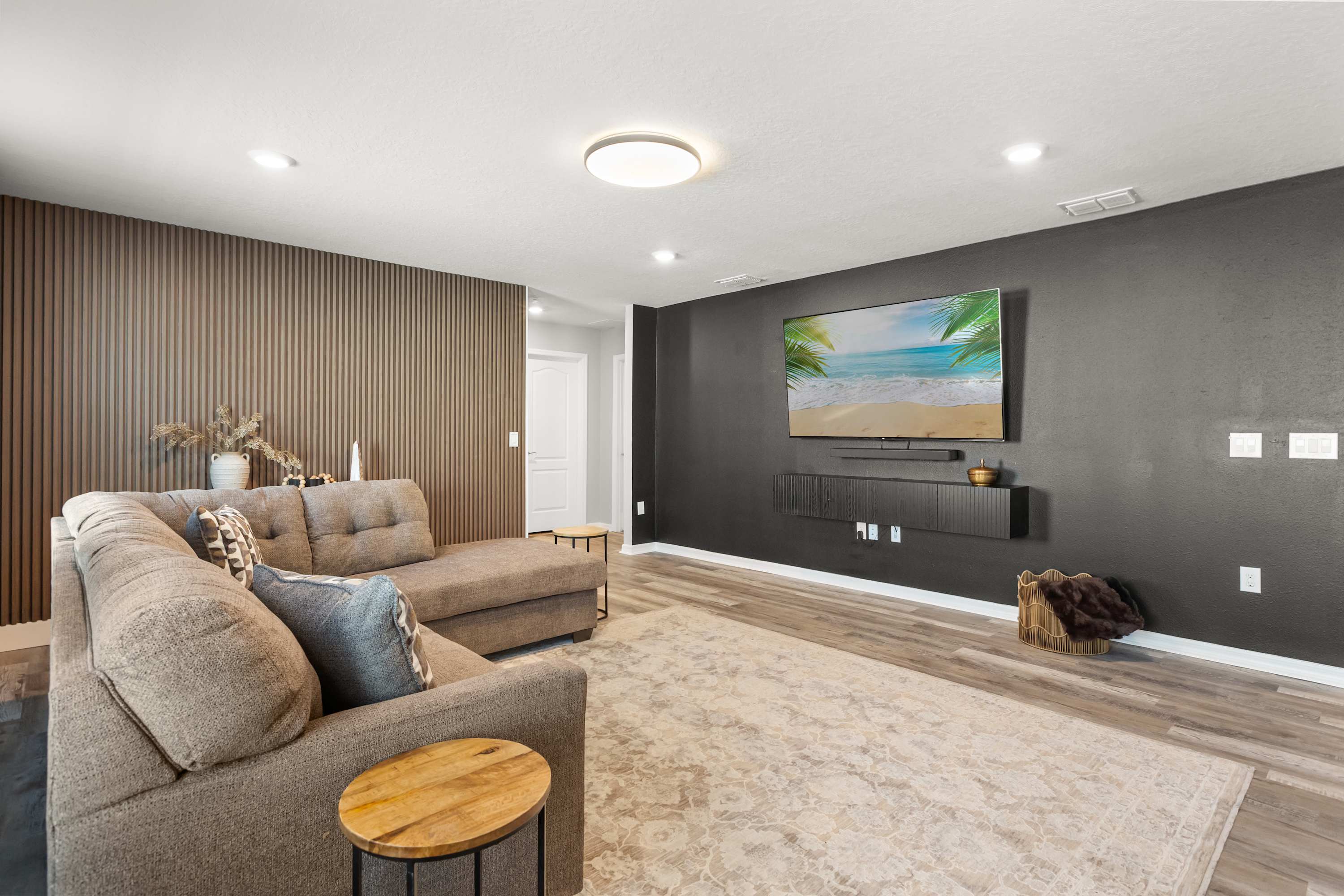 Entertainment room featuring a large L-shaped sectional sofa facing a wall-mounted TV with a floating media console. One wall is accented with vertical wood paneling, while the TV wall is painted dark gray, all on wood-look laminate flooring.