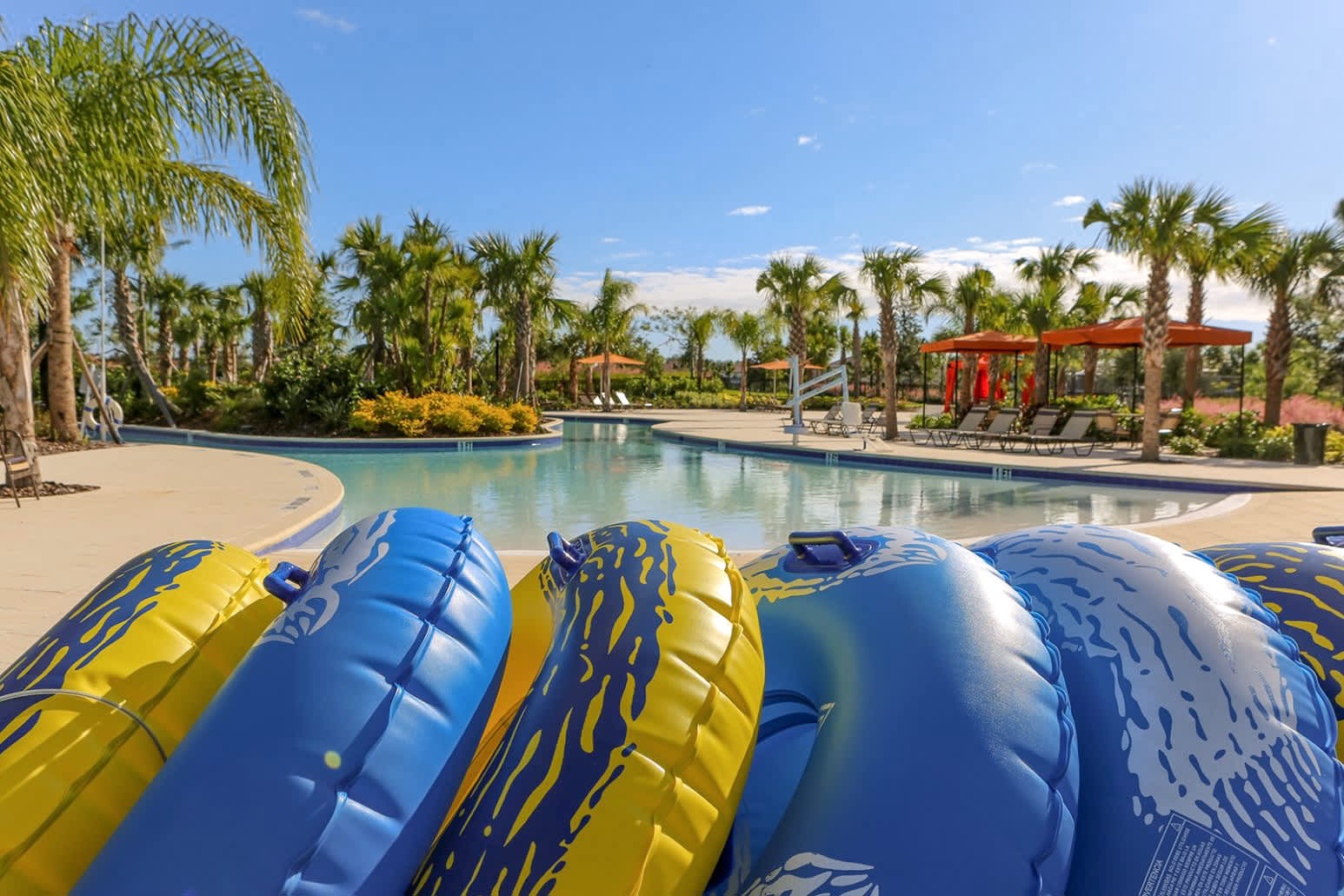 A row of vibrant blue and yellow inflatable tubes rests on the edge of a lazy river, with lush palm trees, sun umbrellas, and distant lounge areas creating a tropical and relaxing atmosphere under a clear sky.