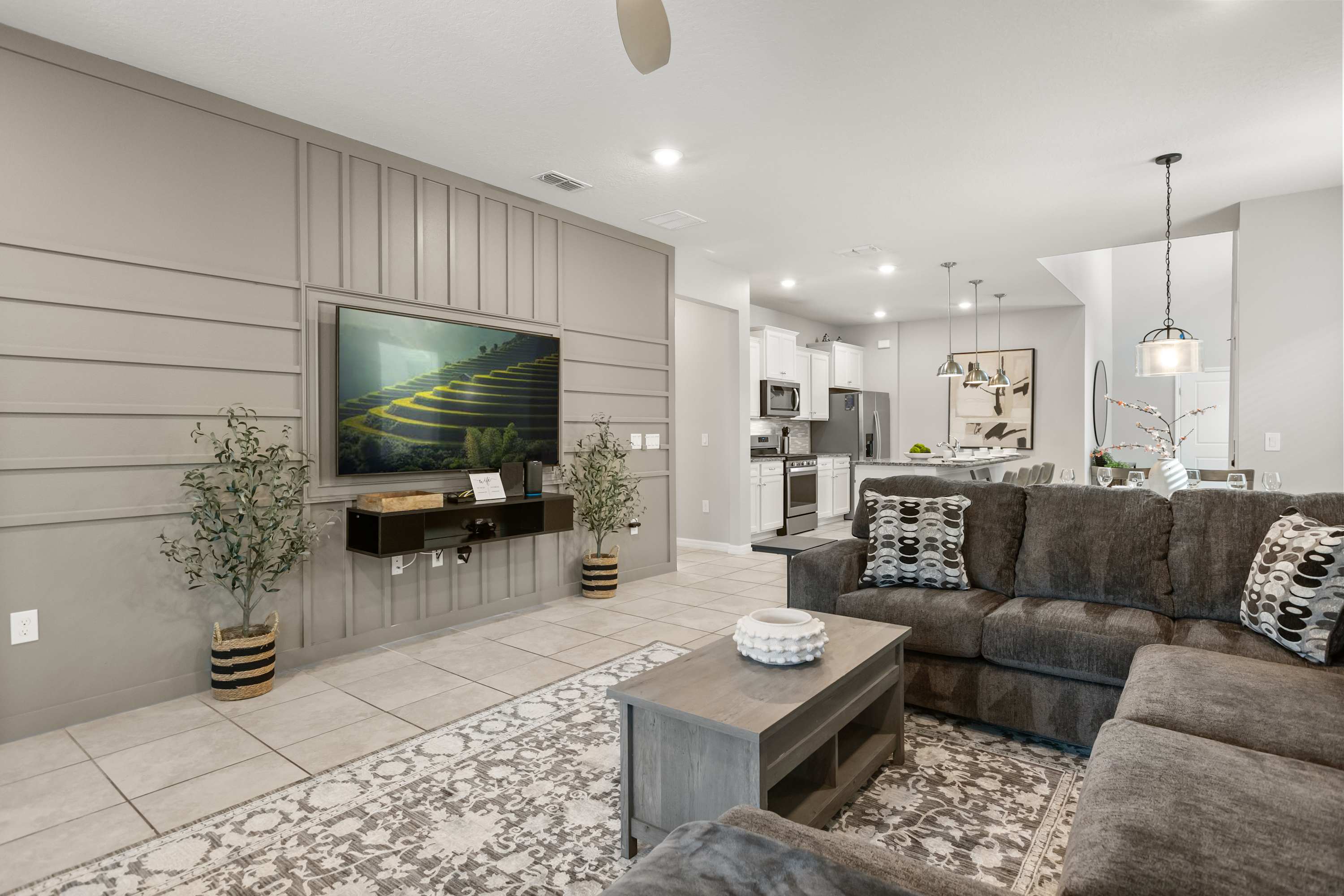 An open-concept living room features a large gray sectional sofa, a wooden coffee table, and a wall-mounted TV against a light gray paneled accent wall. The space flows into a modern kitchen and dining area in the background.