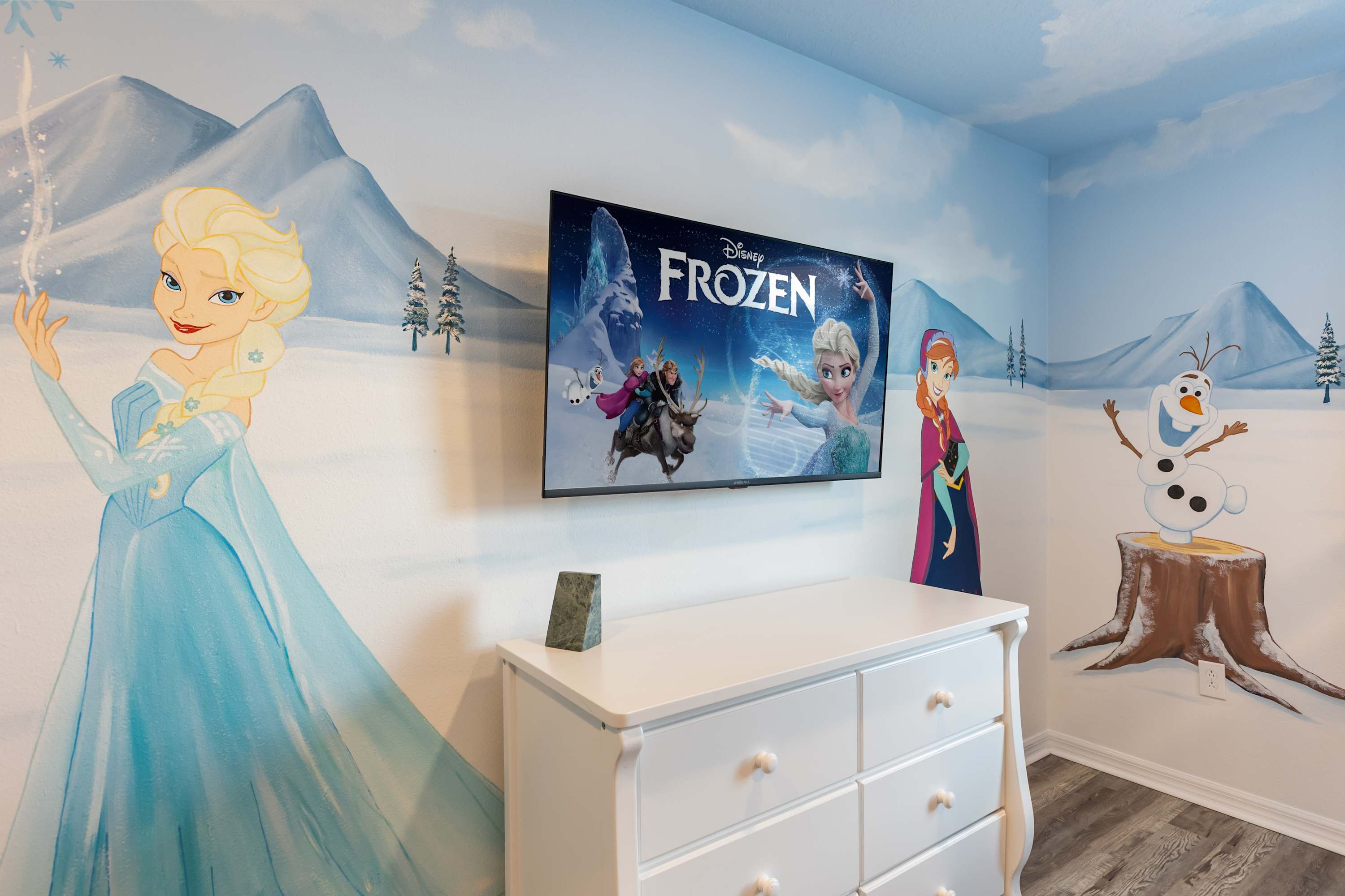 A close-up view of the "Frozen" themed bedroom, showcasing a detailed mural of Elsa on the left wall and Olaf on the right. A wall-mounted TV above a white dresser displays the "Frozen" movie, complementing the playful decor.