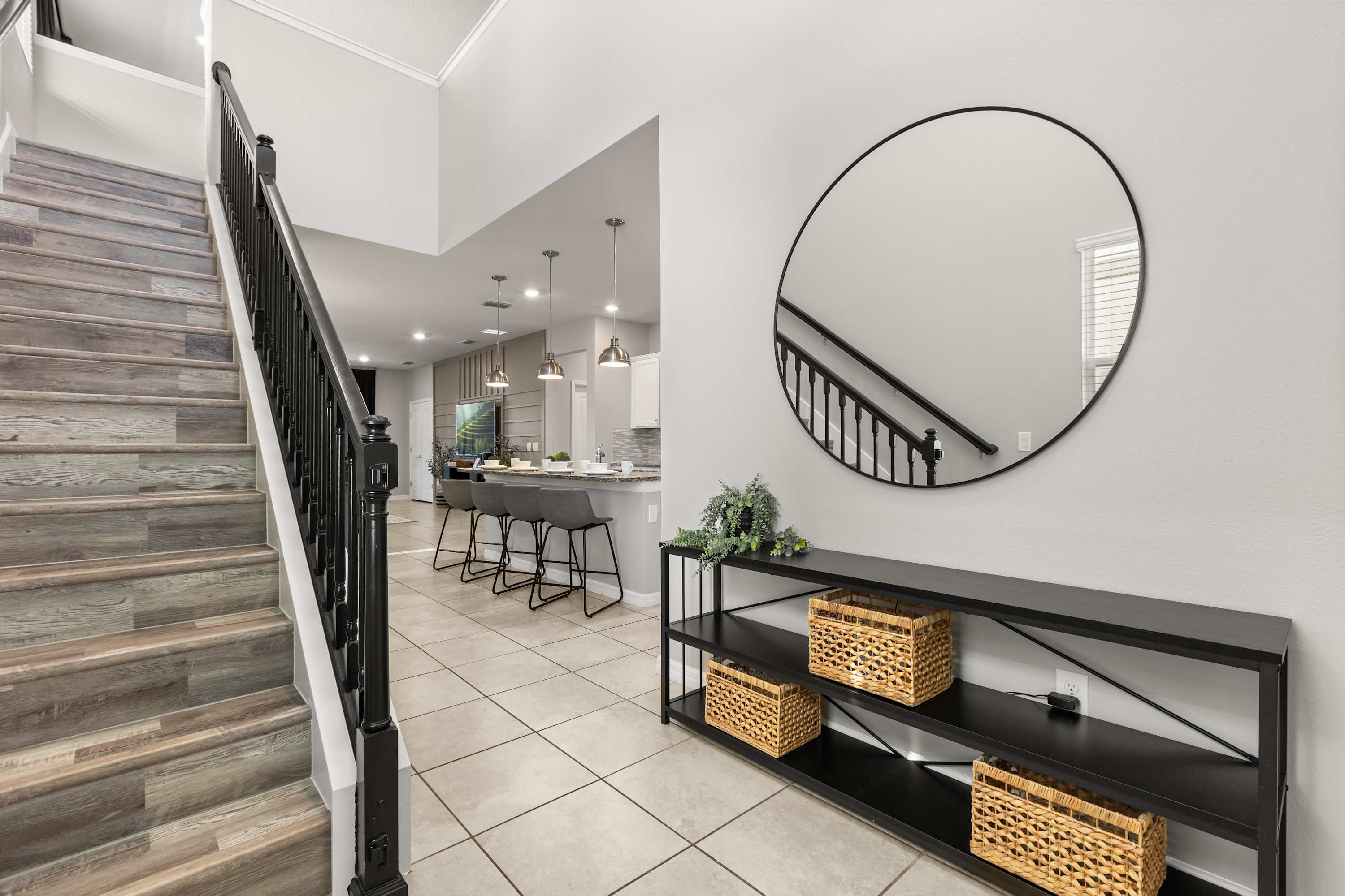 Entryway or foyer area with a modern black console table, wicker baskets, and a large round black-framed mirror. A staircase with wood treads and black railings ascends to the upper floor, with light tile flooring throughout.