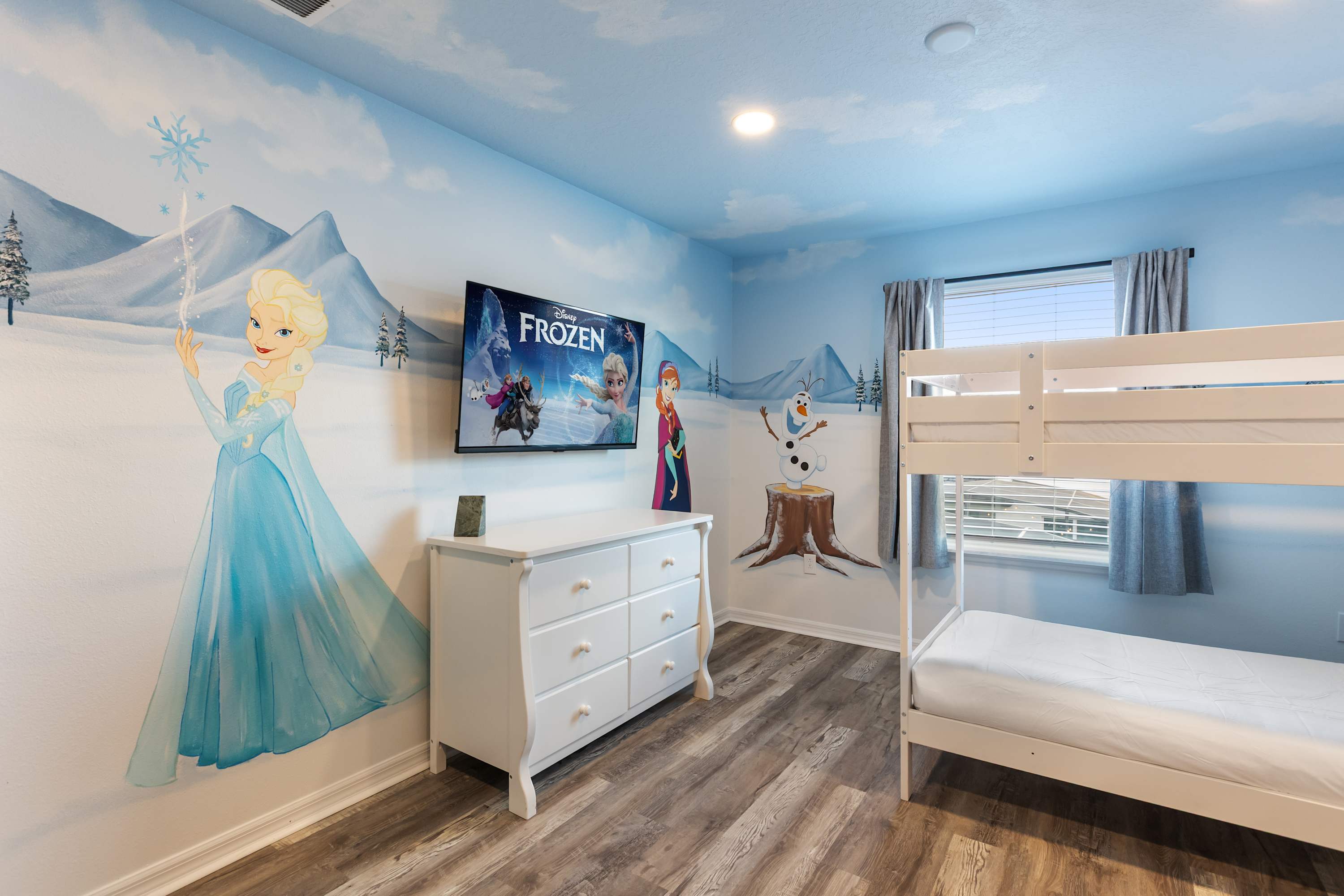 A vibrant kids' bedroom featuring a "Frozen" themed mural on the walls and ceiling, with a white bunk bed on the right. A wall-mounted TV displays the "Frozen" logo above a white dresser, all set on wood-look flooring.