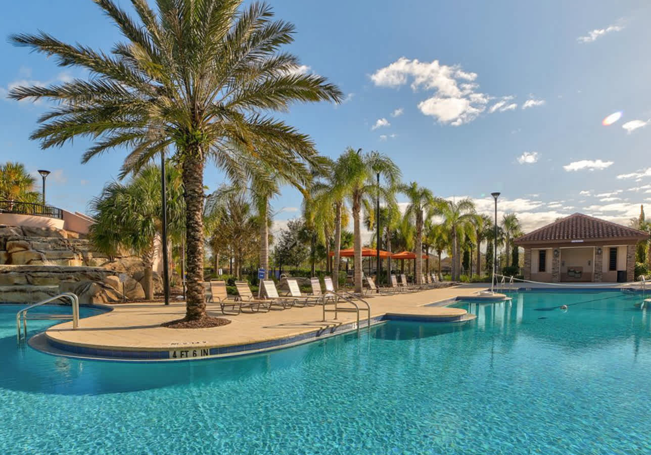 A large, inviting swimming pool with clear blue water is surrounded by a paved deck with numerous lounge chairs, shaded by tall palm trees, with a clubhouse building visible in the background under a bright sky.