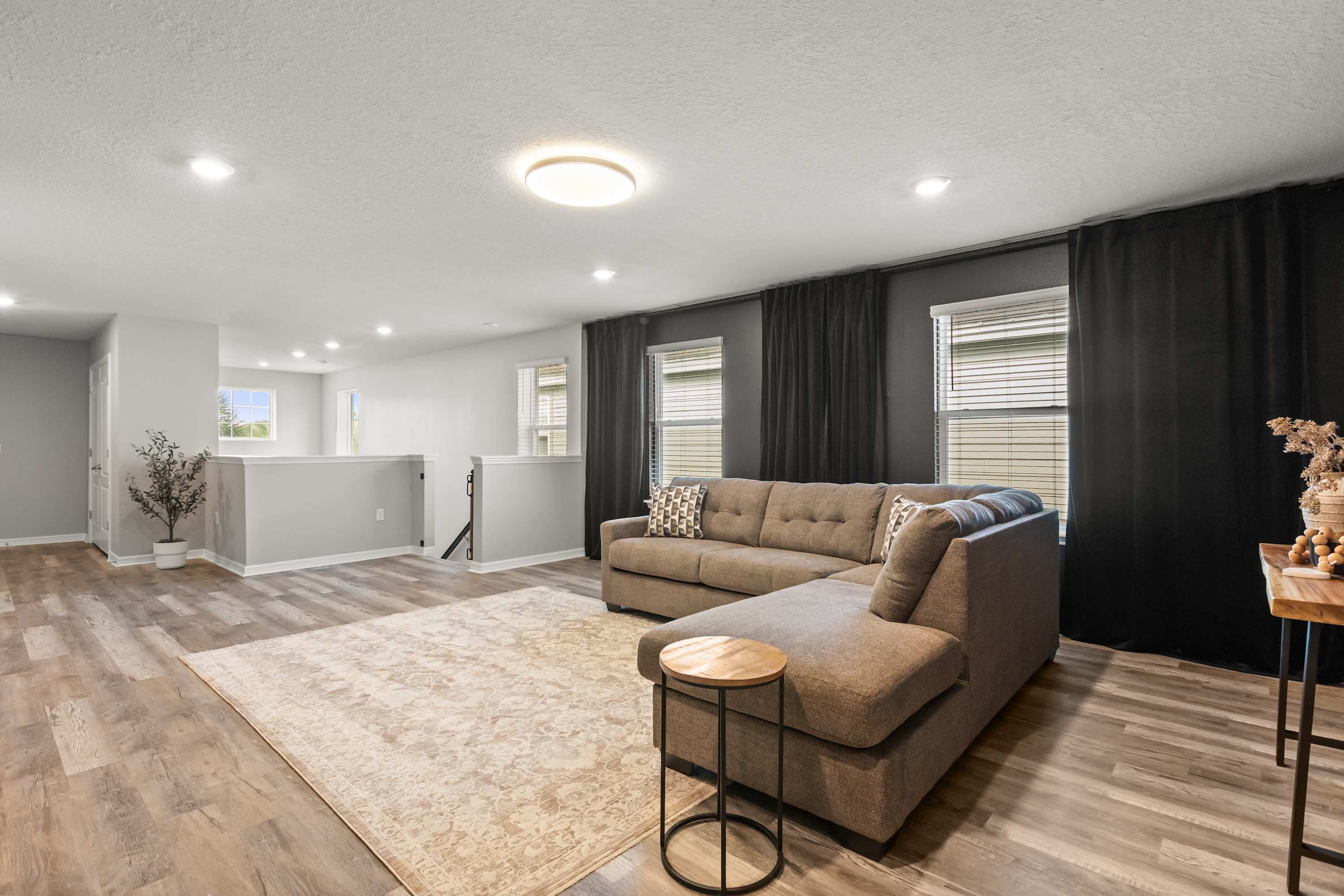 Upper-level entertainment space with a large L-shaped sectional sofa and a light patterned area rug on wood-look laminate flooring. Windows with dark curtains and blinds provide natural light, and a railing overlooks the floor below.