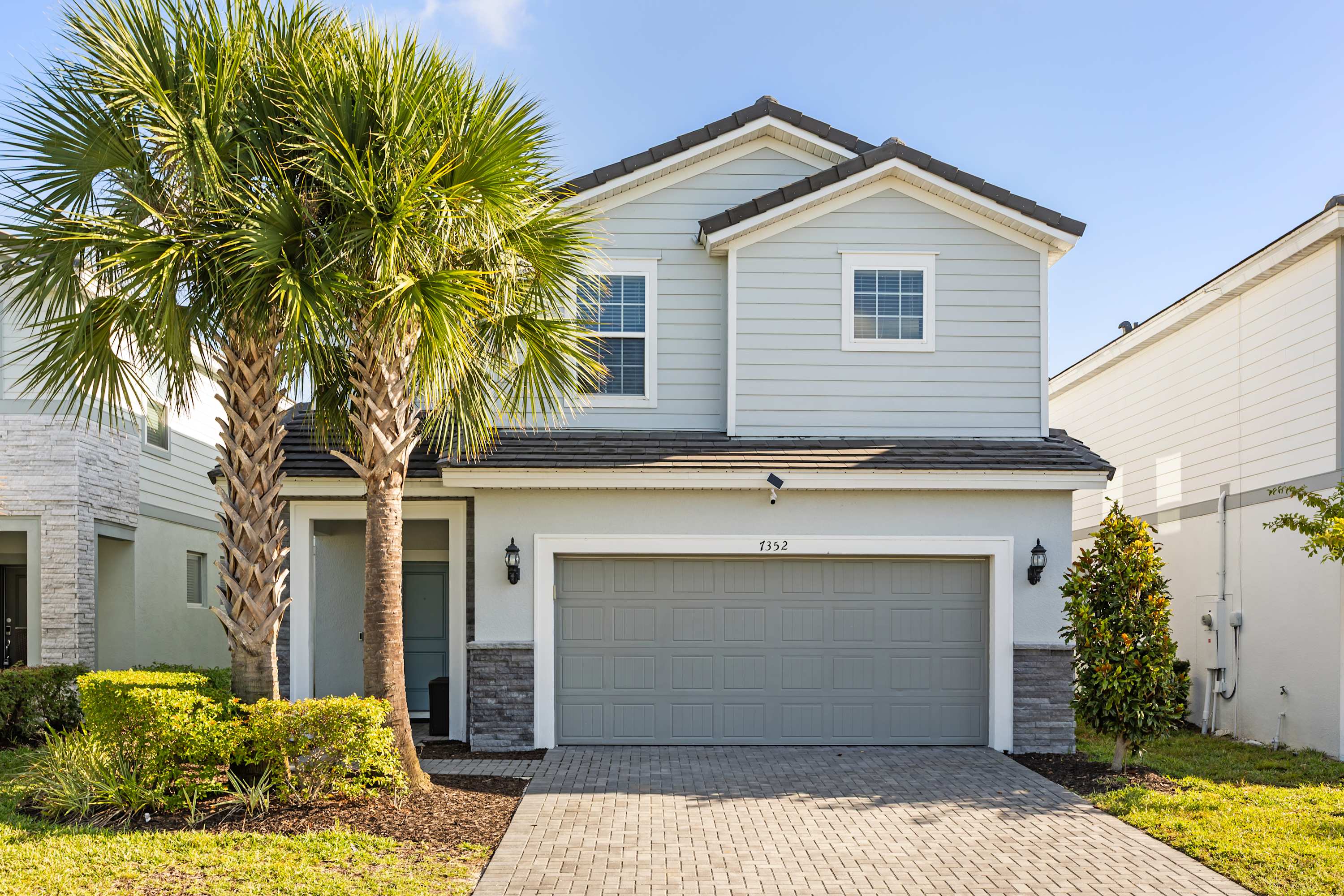 A full frontal view of a modern two-story light blue house with gray siding and stone accents, featuring a two-car garage with a gray door. Lush green grass and two prominent palm trees adorn the front yard, with a paved driveway.