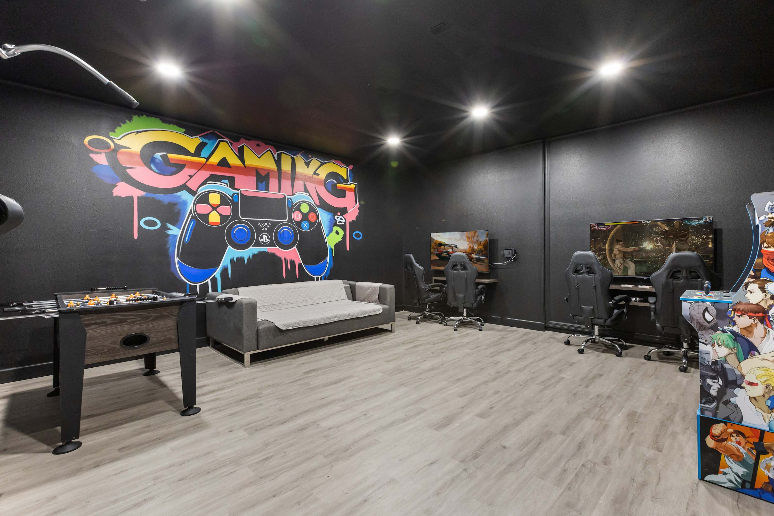 A vibrant game room with black walls features a colorful "GAMING" mural, two gaming stations with chairs, a foosball table, and a gray sofa. The room has light wood-look flooring and bright recessed lighting.