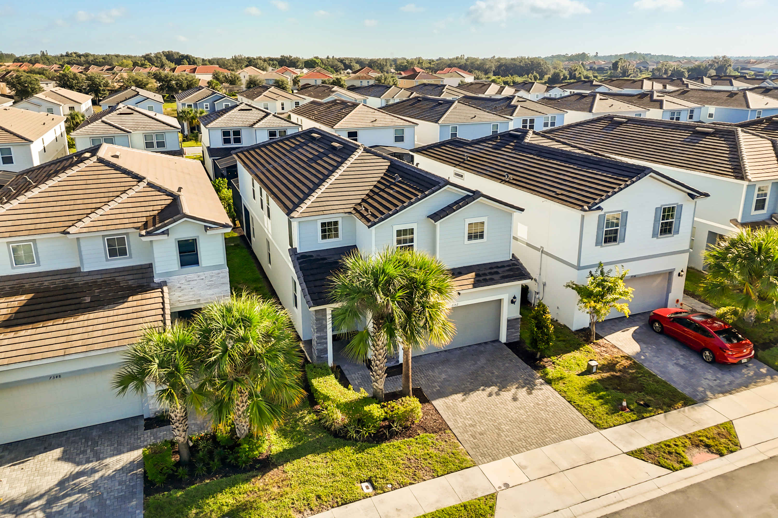 An aerial view showcases a row of modern residential houses with light-colored exteriors and dark roofs, each with a driveway and small front yard, set within a planned community under a clear sky.