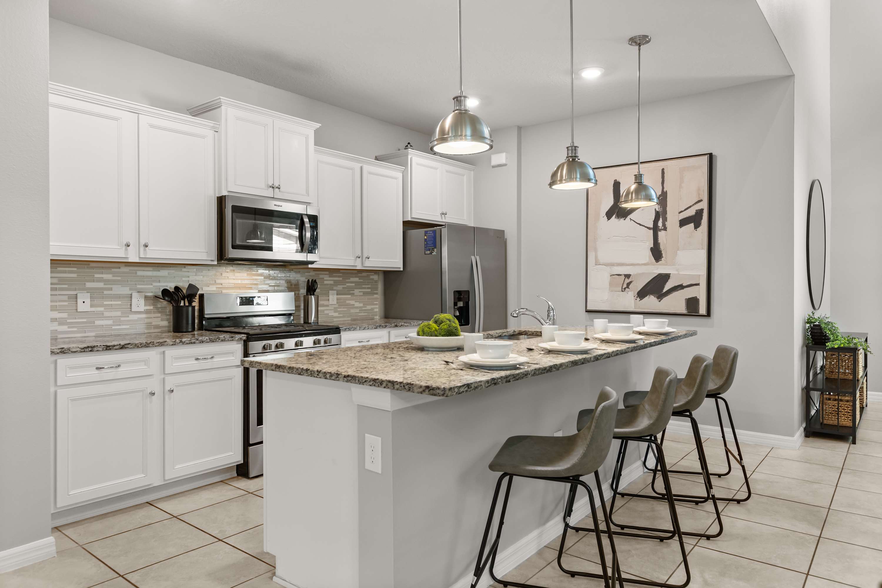 Bright kitchen featuring white shaker cabinets, granite countertops, and stainless steel appliances, including a gas range and microwave. A large island with four gray bar stools provides additional seating, illuminated by three modern pendant lights.