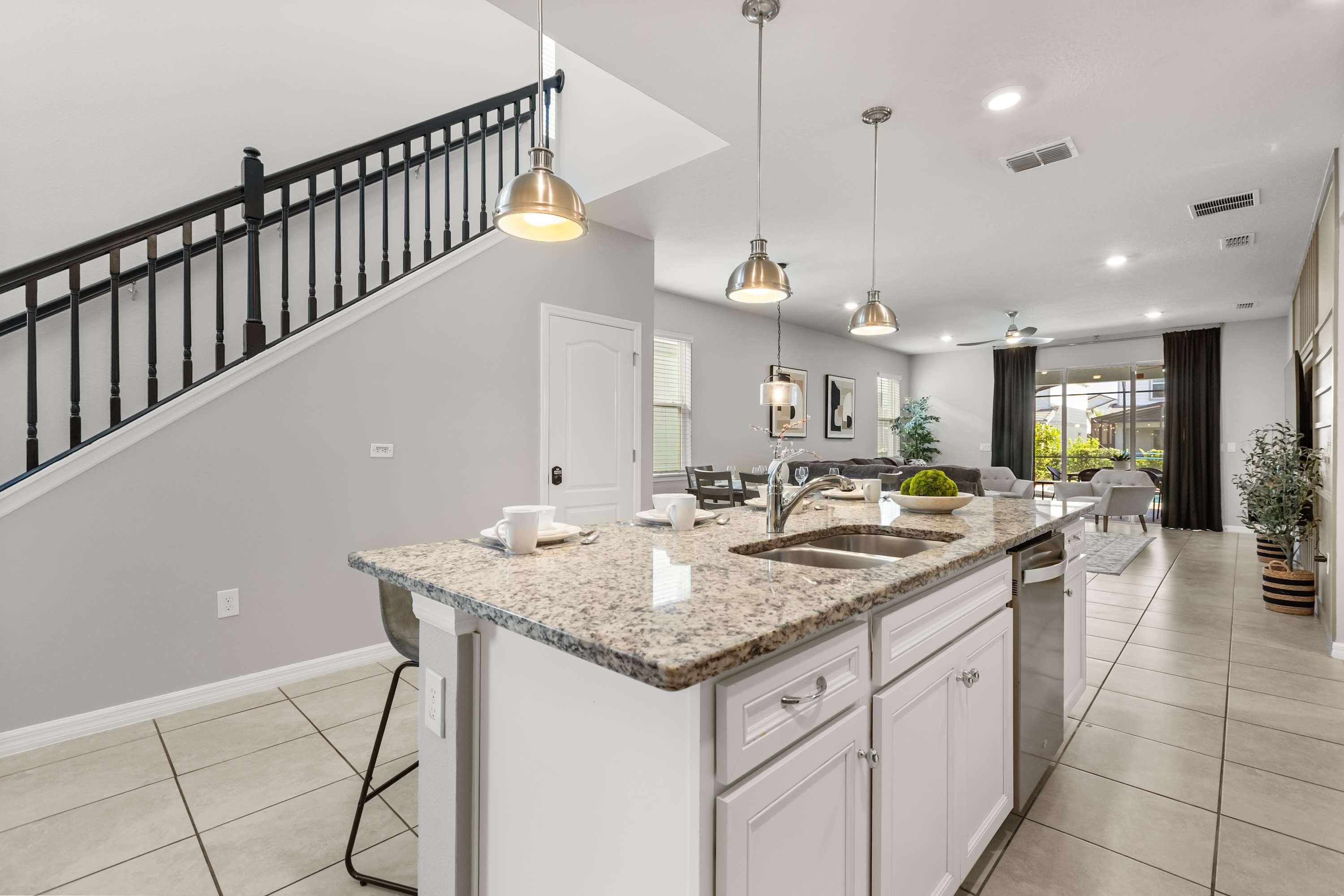 View of the kitchen island with a sink and three pendant lights, showcasing granite countertops and white cabinets. A dark wood staircase with black railings is visible in the background, leading upstairs.