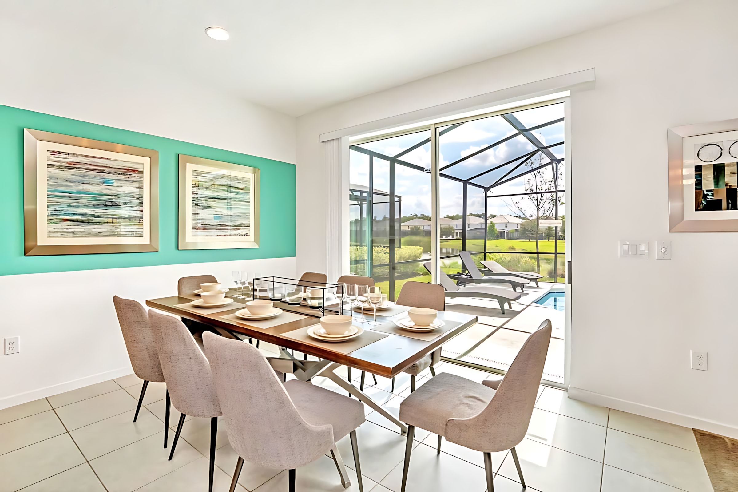 Modern dining room with a long wooden table set for eight, light gray upholstered chairs, and a vibrant teal accent wall with abstract art, overlooking a screened pool area through large glass doors.