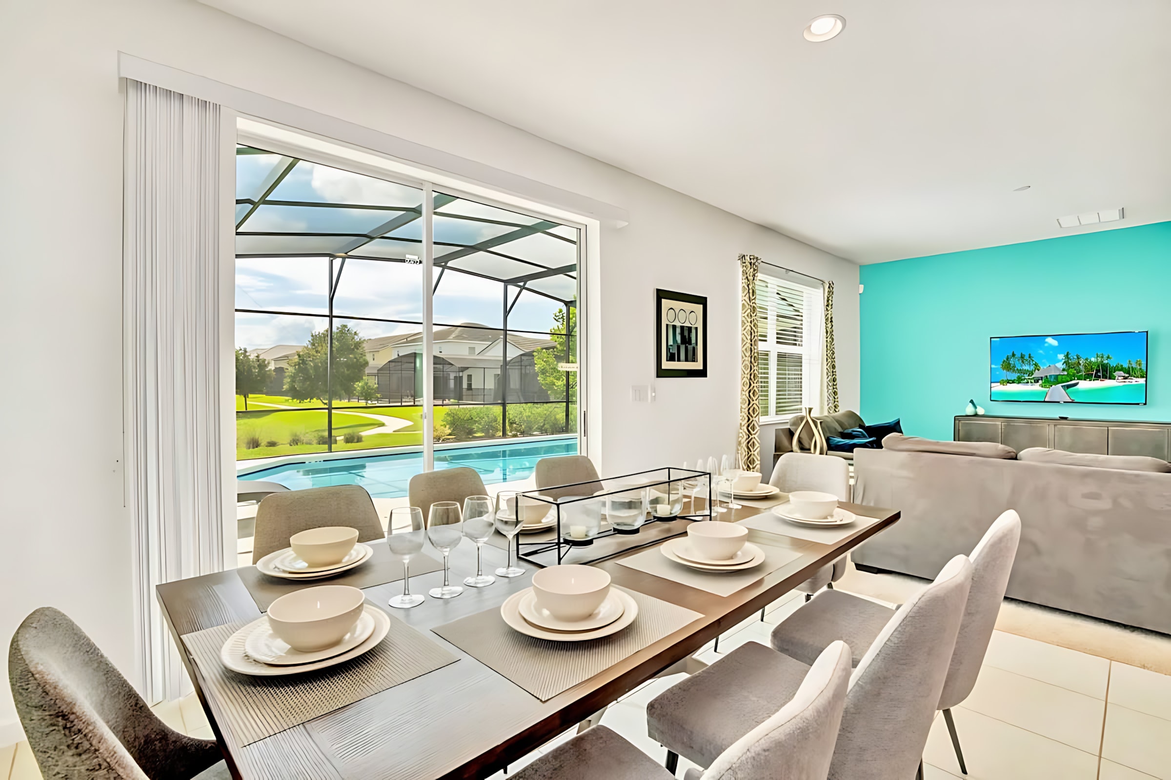 A modern dining area features a long dark wood table set with white dishes and glassware, surrounded by light grey upholstered chairs. A large window offers a view of the screened-in pool and backyard.