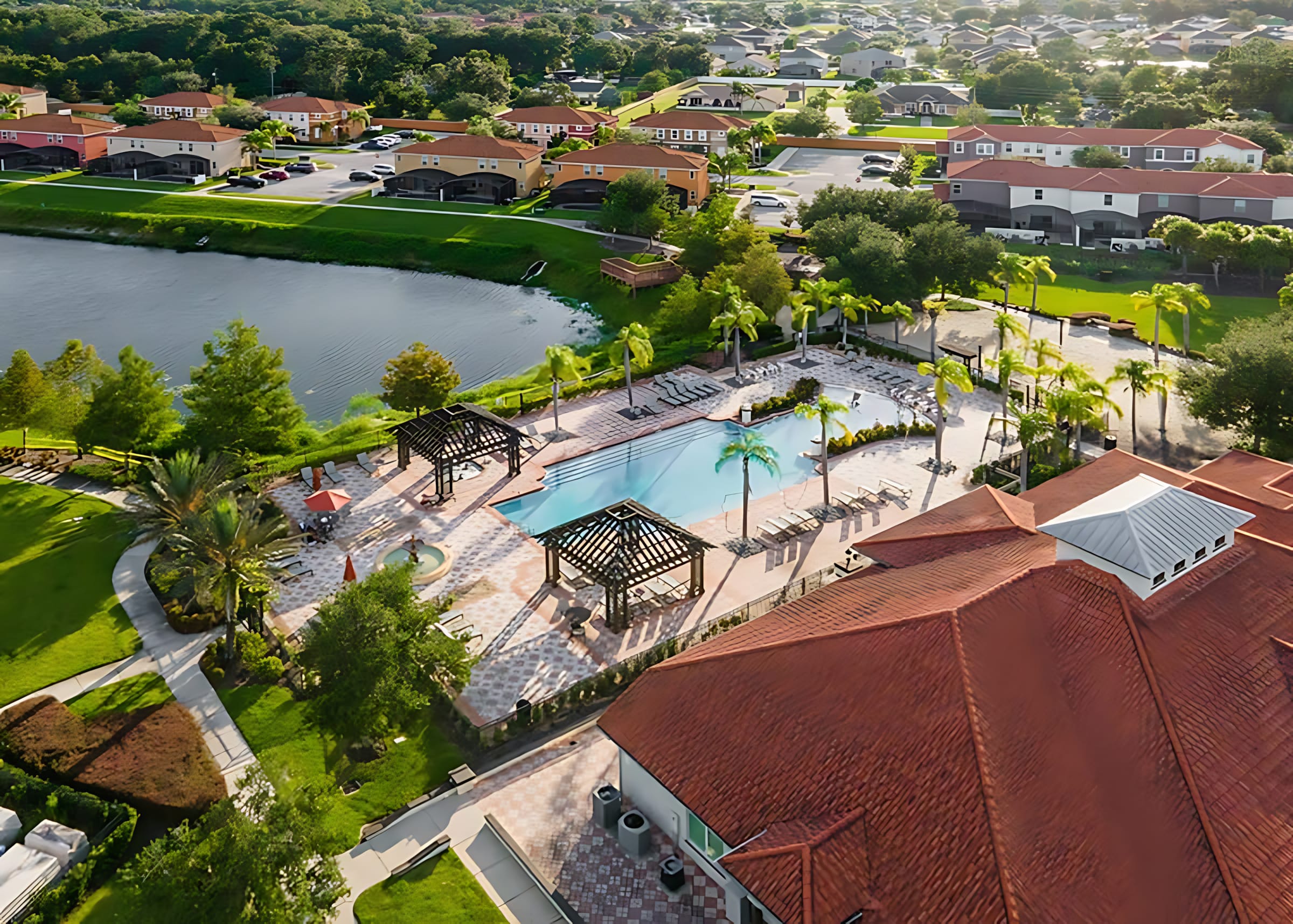 An aerial view showcasing a large communal pool area with two gazebos and numerous lounge chairs, surrounded by patterned pavers and palm trees. The pool is adjacent to a lake, with residential homes and lush green landscaping visible under a bright sky.