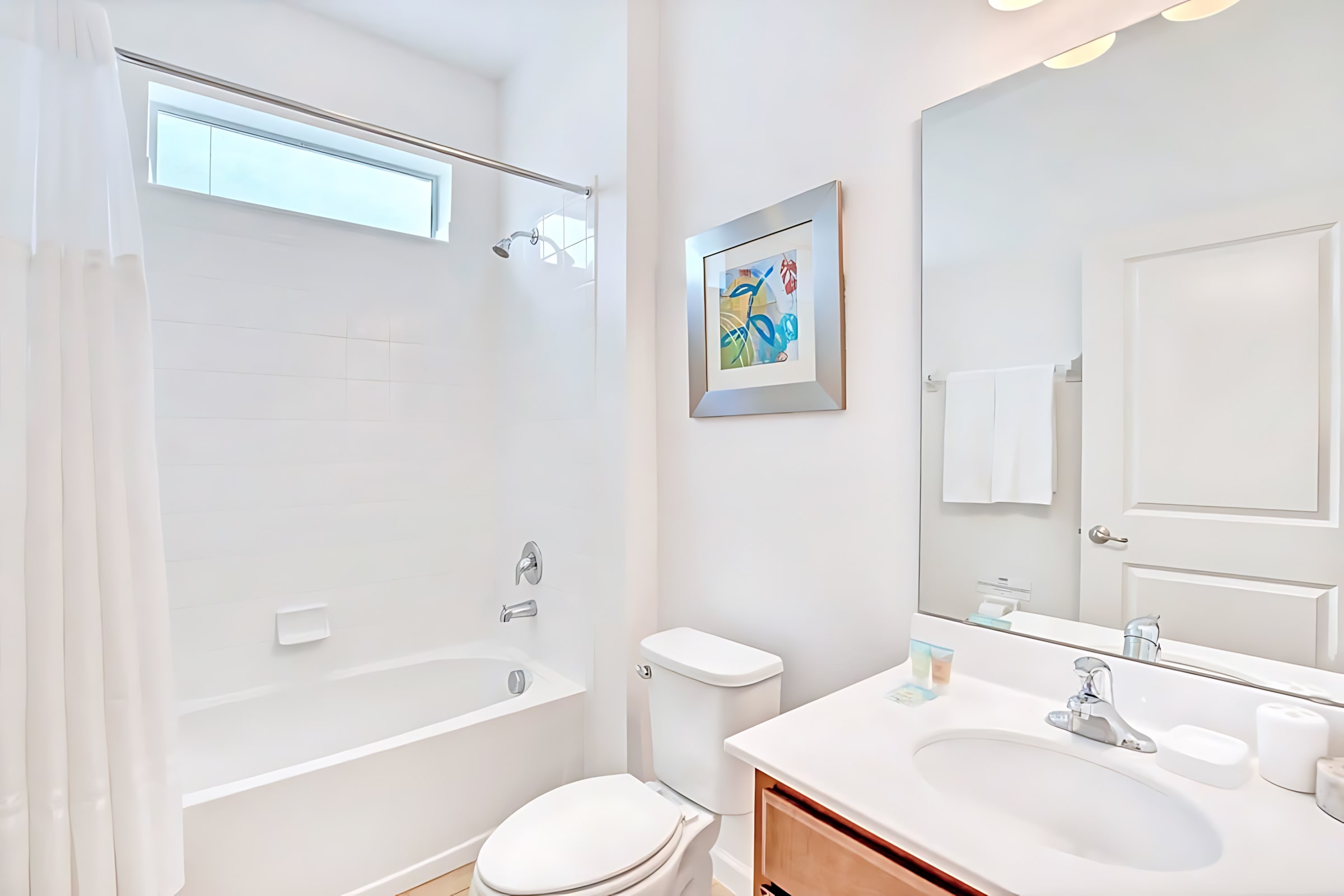 Clean white bathroom featuring a bathtub with a shower, white subway tile surround, and a small horizontal window above. A white shower curtain is pulled back, and a toilet is visible next to the tub.