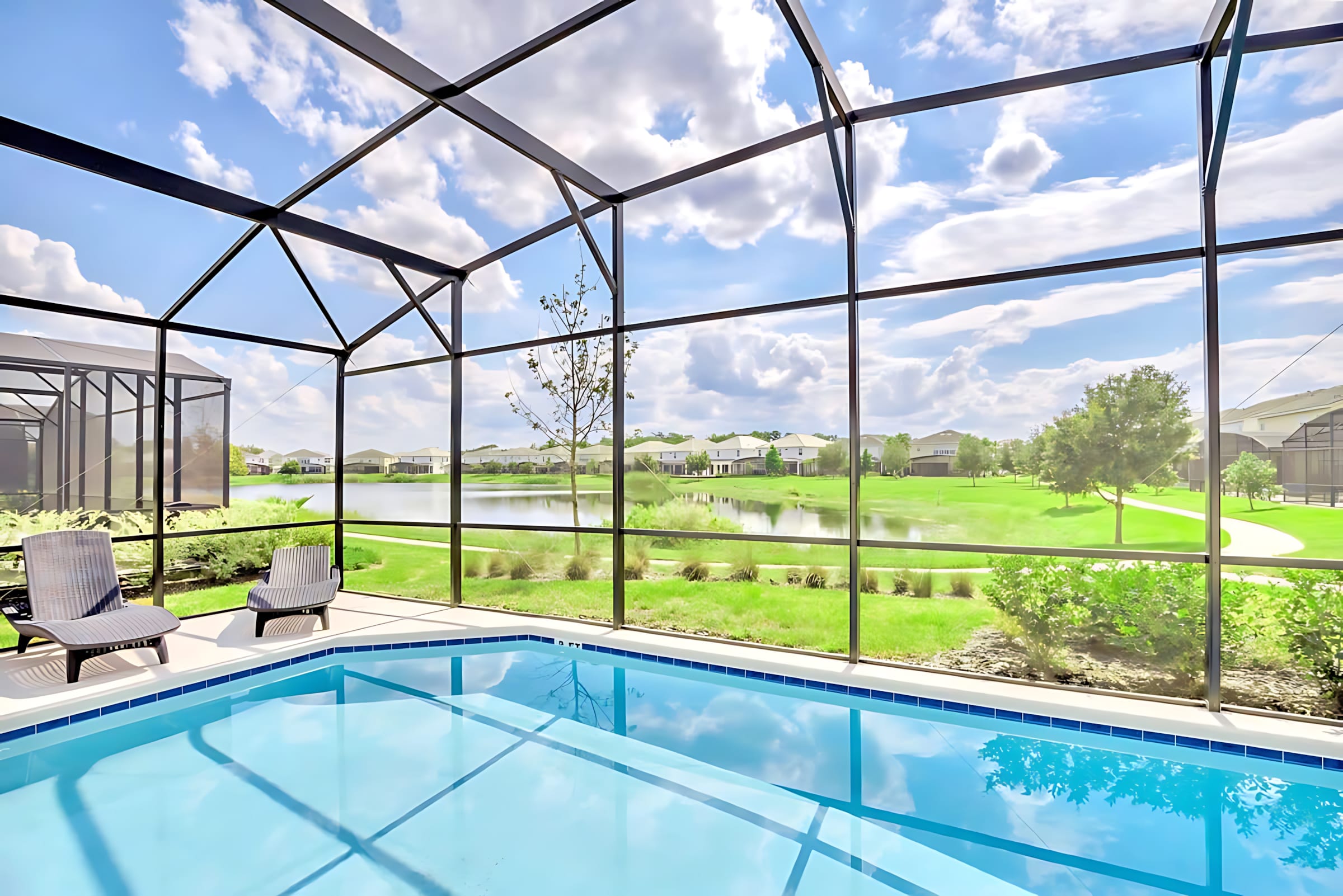 A close-up view of the screened-in private pool, showcasing its light blue water and the surrounding patio area with two lounge chairs. The background reveals a serene lake and residential homes under a bright sky.