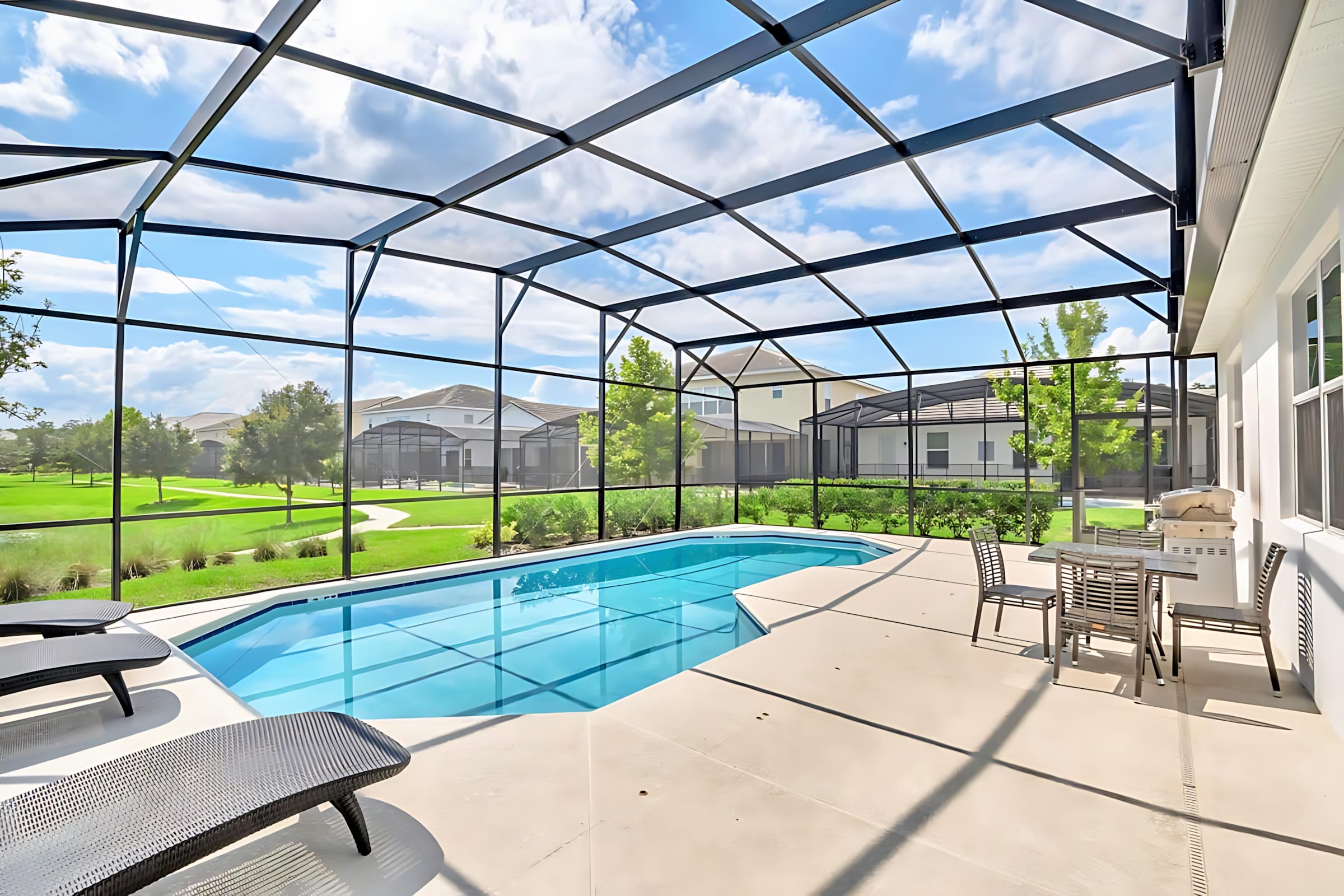 A screened-in private pool area features a light blue swimming pool, two dark lounge chairs, and a small dining set with a BBQ grill. The enclosure offers views of a grassy area and distant houses under a blue sky.
