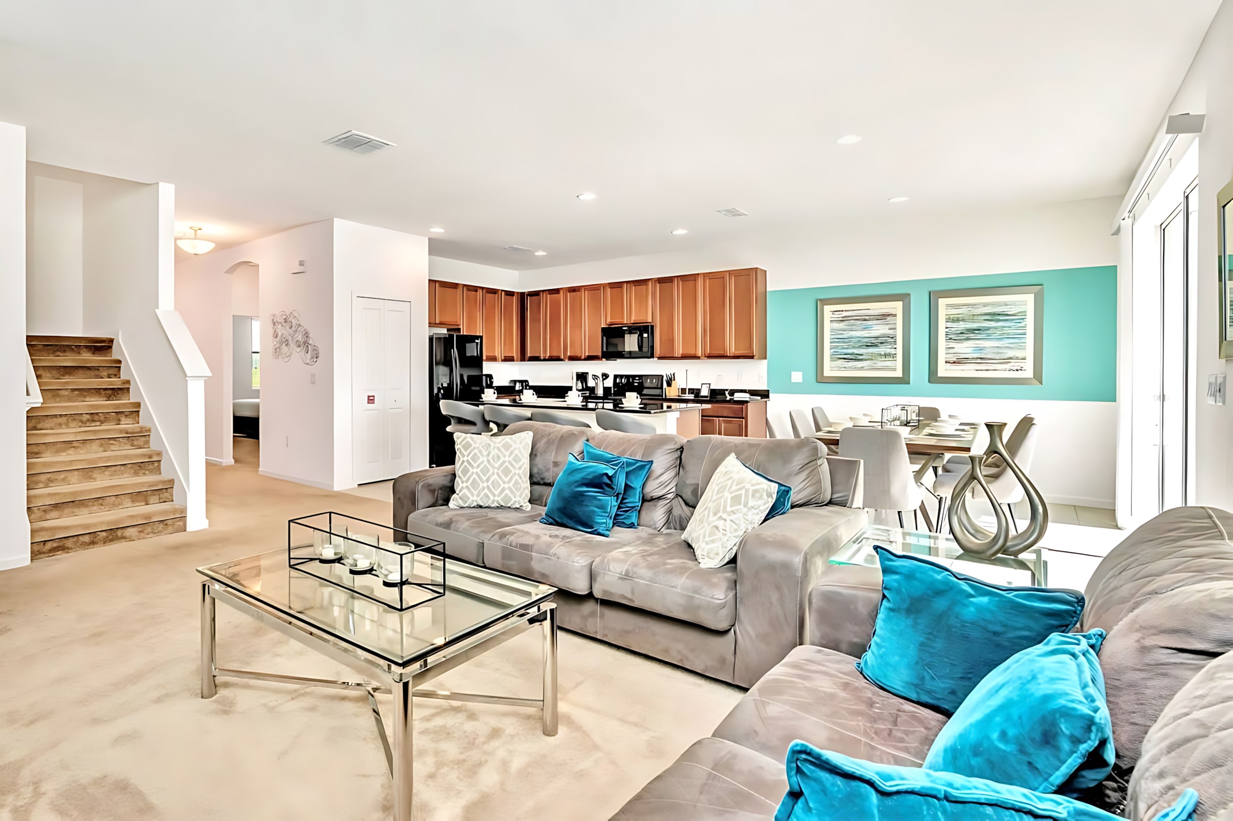 An open-concept living room features two large grey sofas with blue and white throw pillows, a glass coffee table, and a view towards the kitchen and dining area. A carpeted staircase is visible on the left.