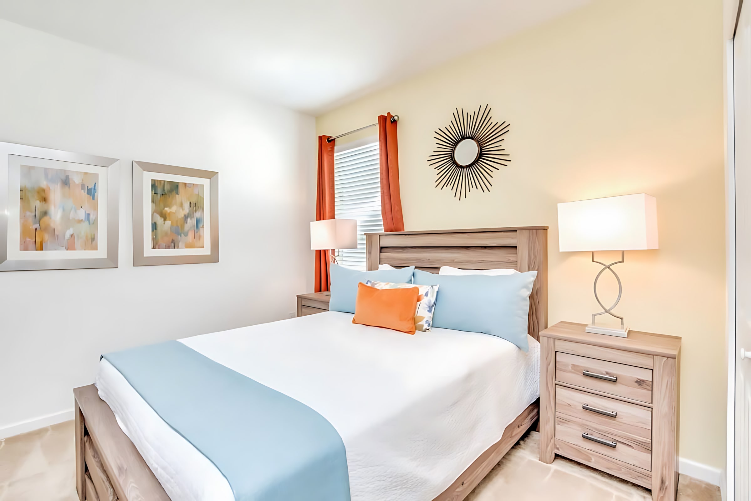 Cozy bedroom with a queen bed featuring a wooden headboard, white bedding with a light blue runner, and an orange accent pillow. The walls are light yellow, and a sunburst mirror hangs above a nightstand with a lamp.