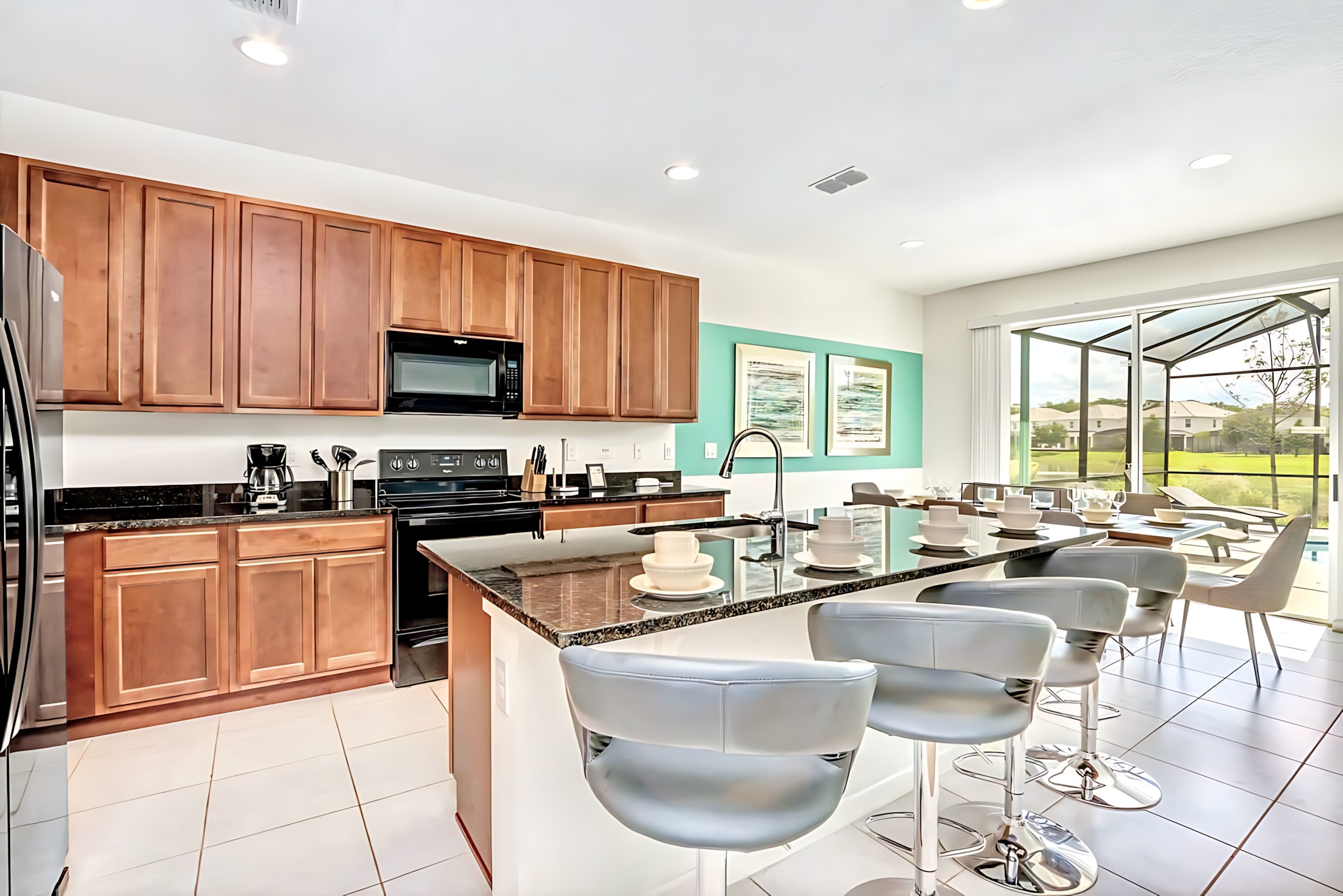 Modern kitchen with dark wood cabinets, black appliances, and dark granite countertops, featuring a large island with a double sink and three gray bar stools, connecting to the dining area and pool view.
