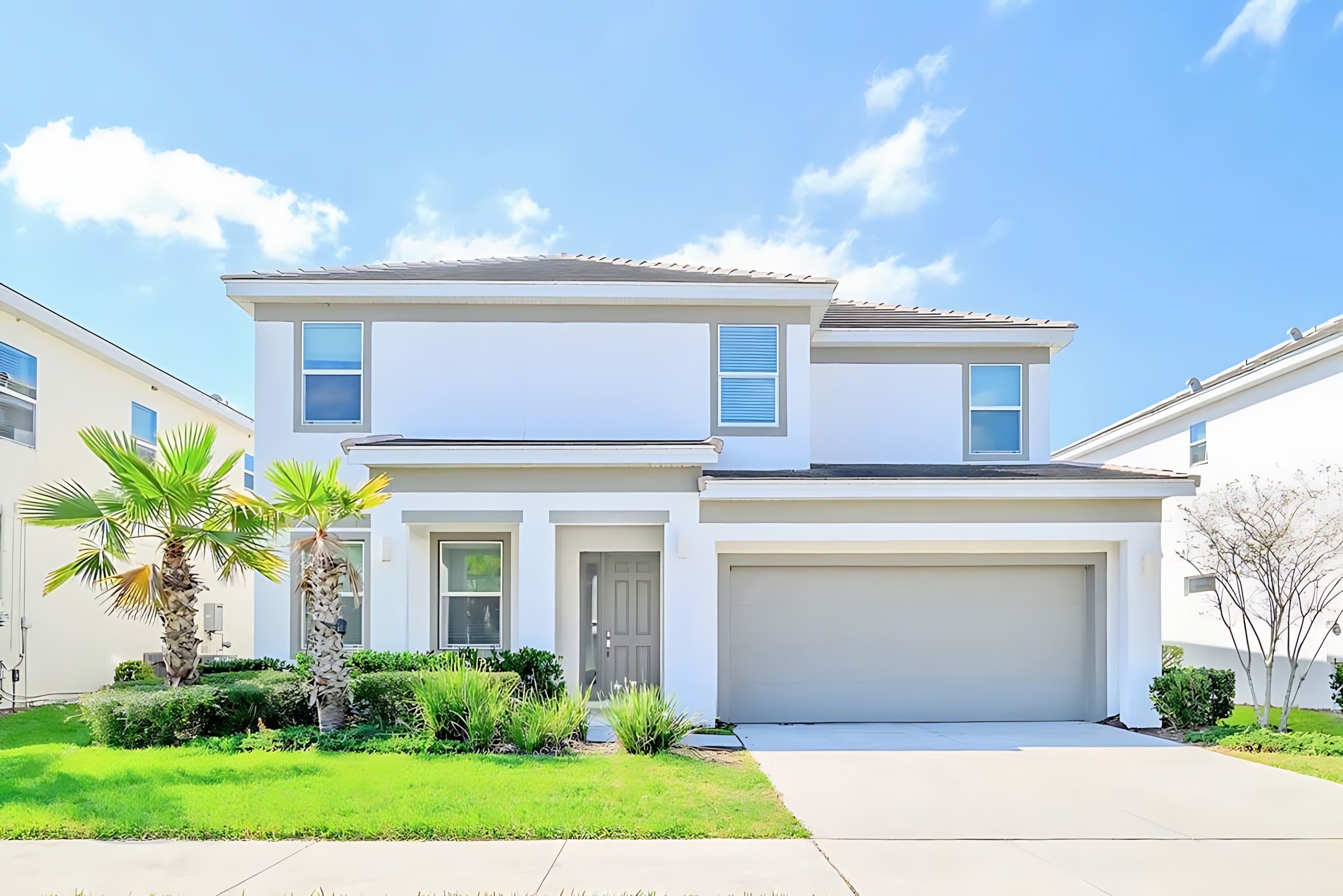 The front exterior of a modern two-story house with white siding and grey trim, featuring a two-car garage and a neatly landscaped front yard. Palm trees and green bushes adorn the concrete driveway leading to the entrance.