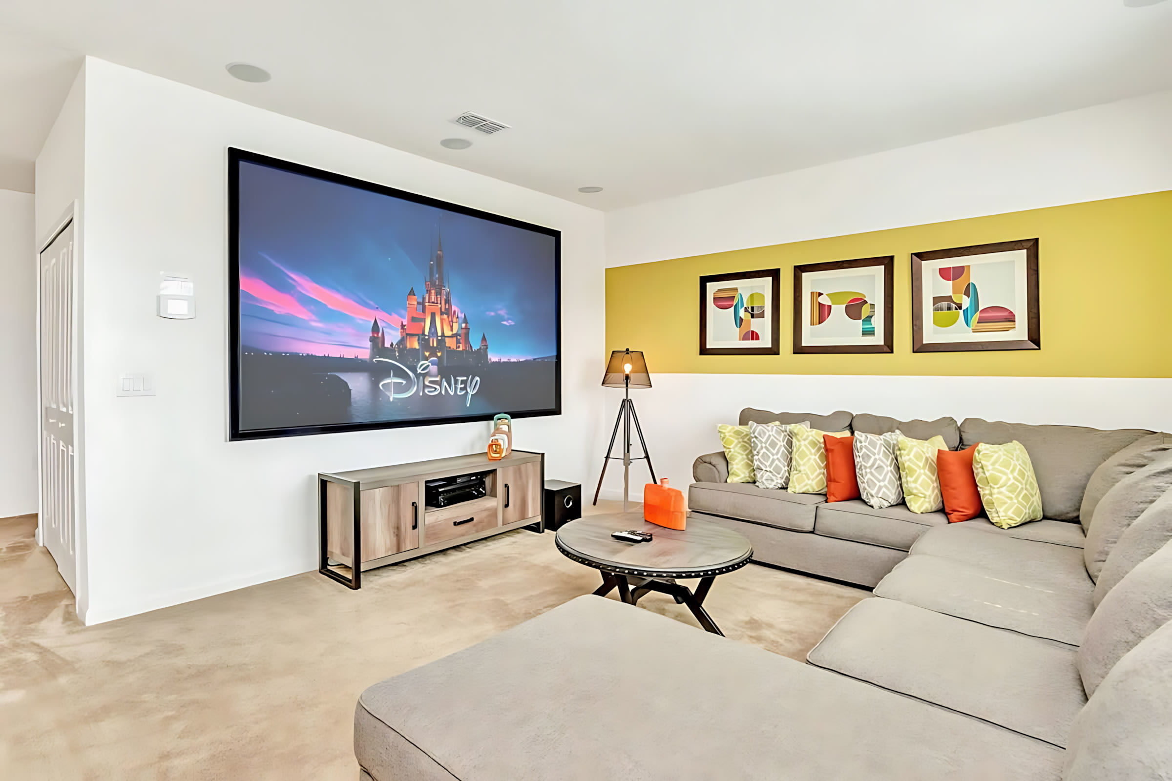 A dedicated media room features a large projection screen displaying a Disney castle, a grey sectional sofa with colorful throw pillows, and a round coffee table. An accent wall with yellow and framed artwork adds a playful touch.