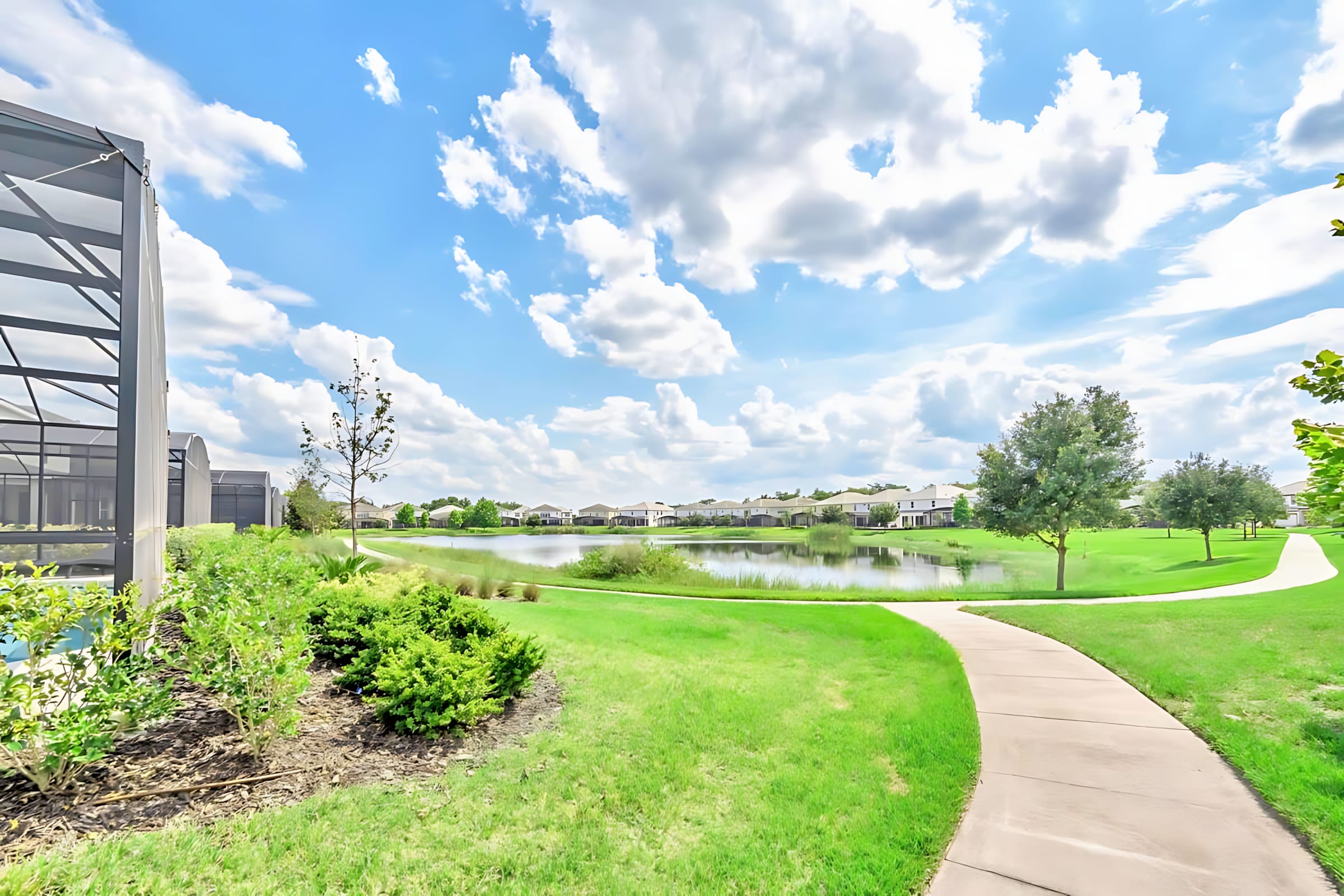 Scenic outdoor view featuring a winding paved pathway alongside a lush green lawn, leading towards a tranquil lake. Residential homes are visible in the distance under a bright blue sky with fluffy white clouds.