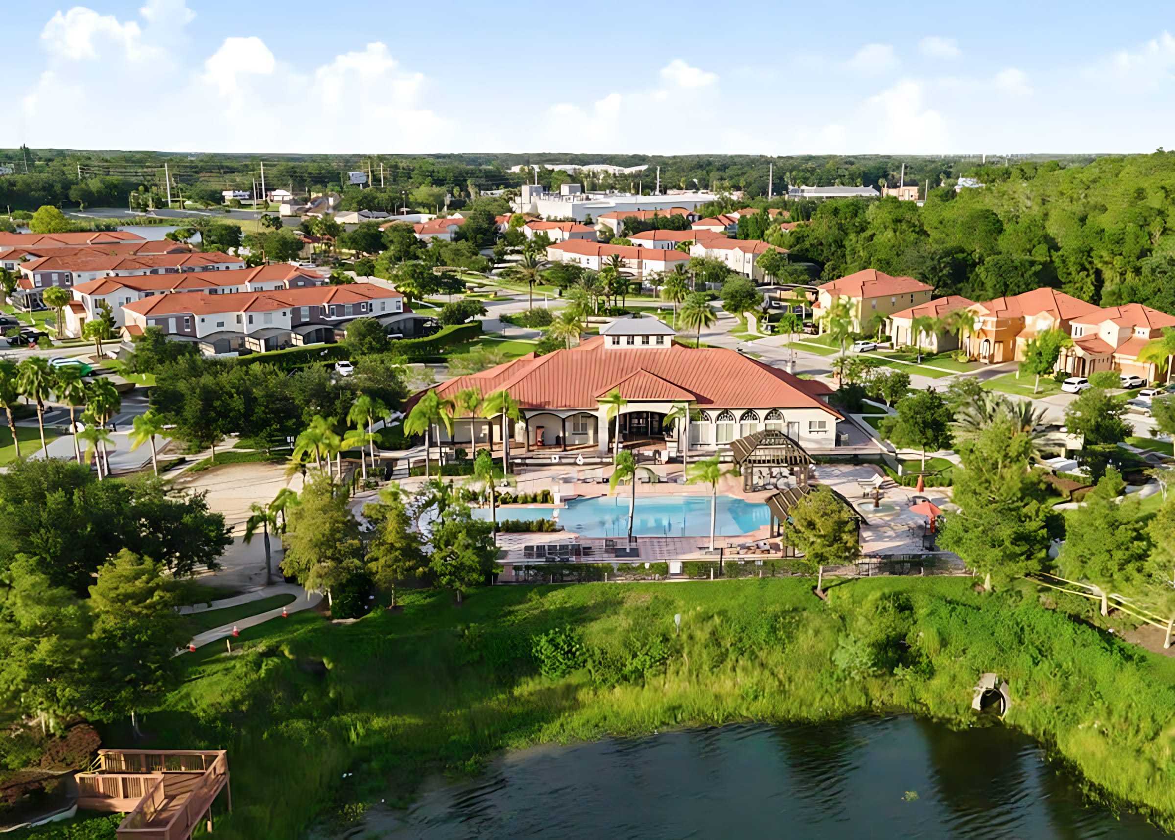 A wide aerial shot of a resort community featuring a large clubhouse with a red tile roof and an expansive communal pool area with gazebos. The property is surrounded by numerous residential homes, lush greenery, and a serene lake under a partly cloudy sky.
