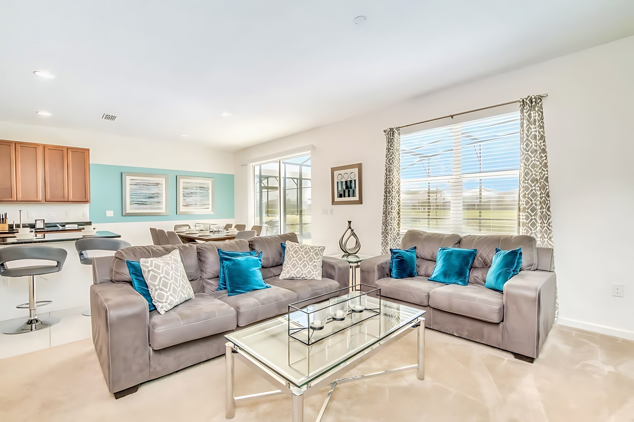 A spacious open-concept living room features two grey sofas with blue and white throw pillows, and a glass coffee table. The room flows into the kitchen and dining area, with a large window offering a glimpse of the outdoor pool.