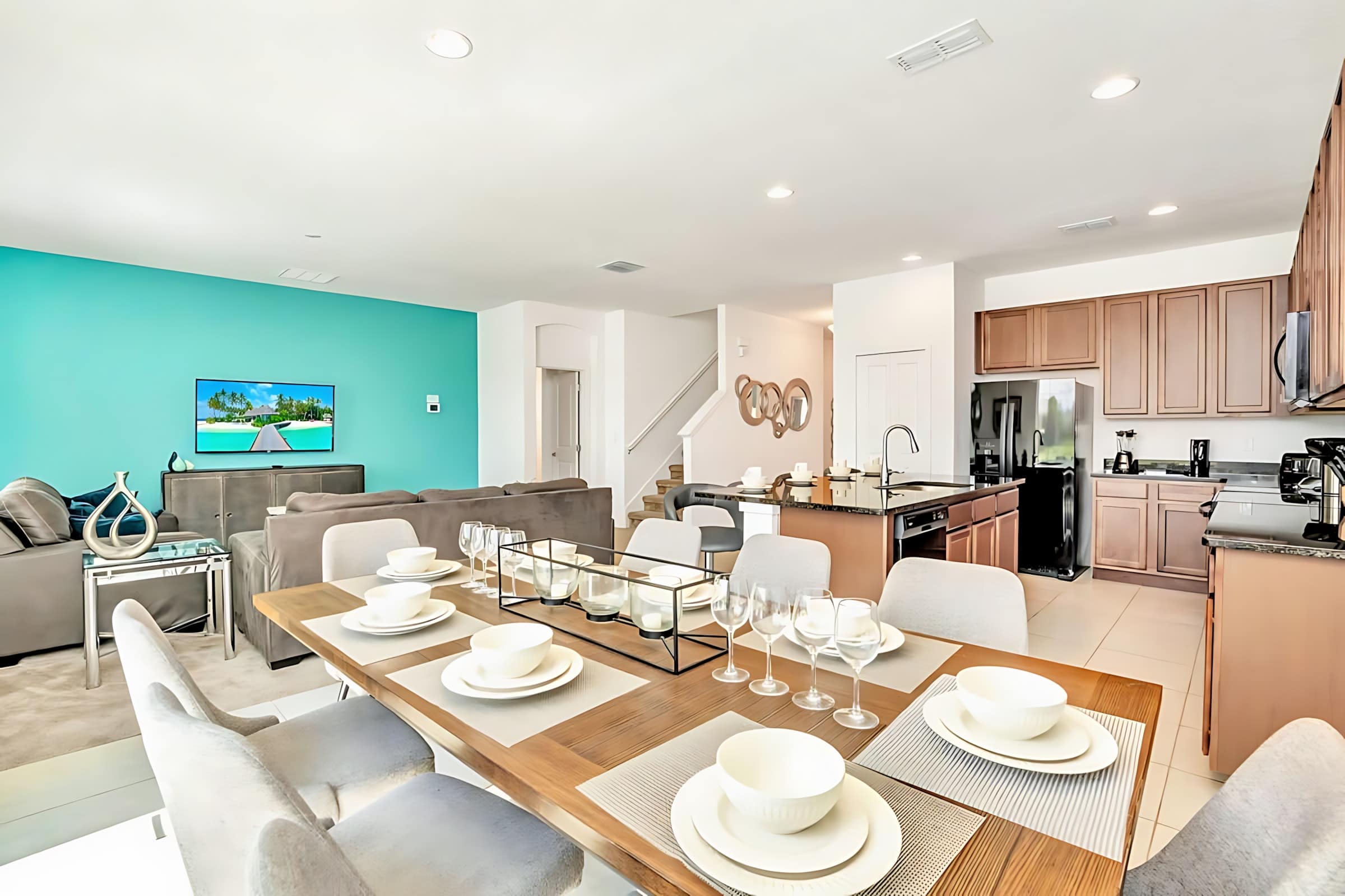 Open-concept living space featuring a dining table set for eight, a comfortable gray sectional sofa facing a wall-mounted TV, and a modern kitchen with dark wood cabinets and granite countertops in the background.