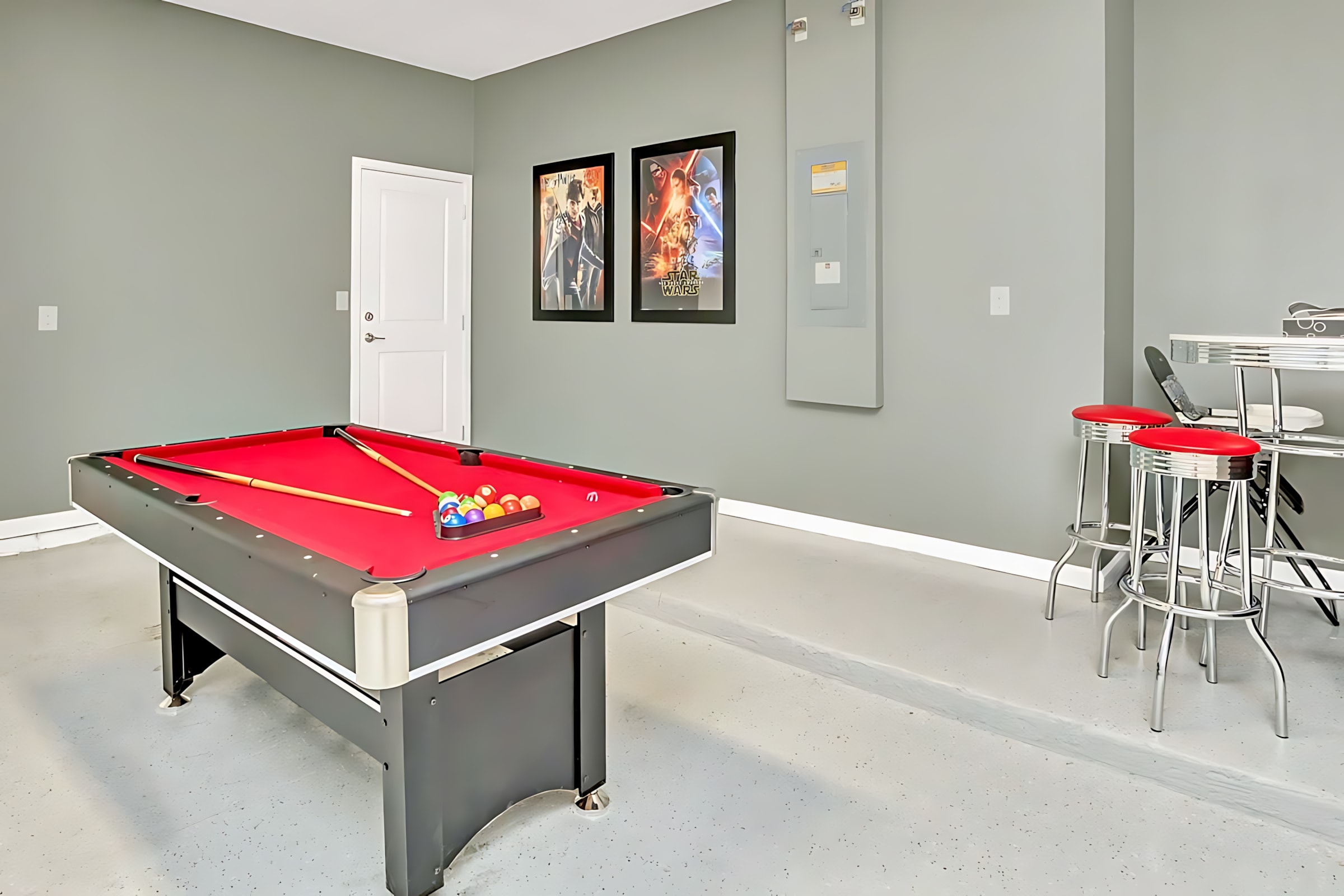 A game room features a red-felted pool table with cues and balls, and a high-top bar table with red stools. The grey walls are decorated with movie posters, creating a fun entertainment space.