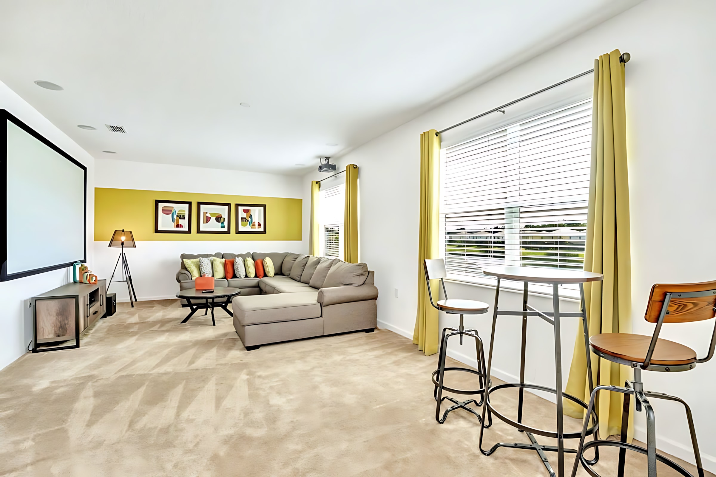 Spacious media room with a large projector screen, a comfortable gray sectional sofa, a round coffee table, and a yellow accent wall with framed artwork, featuring light carpet and bar-height seating by the window.