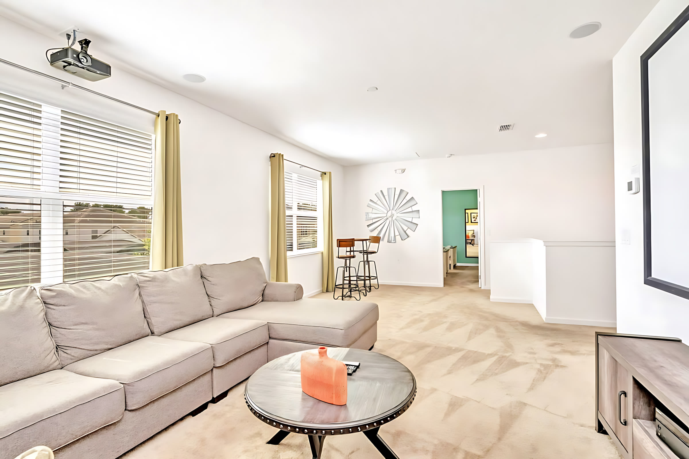Bright media room with a large gray sectional sofa, a round metal coffee table, and a large decorative metal windmill art piece on the wall, offering a view towards a hallway and another room.