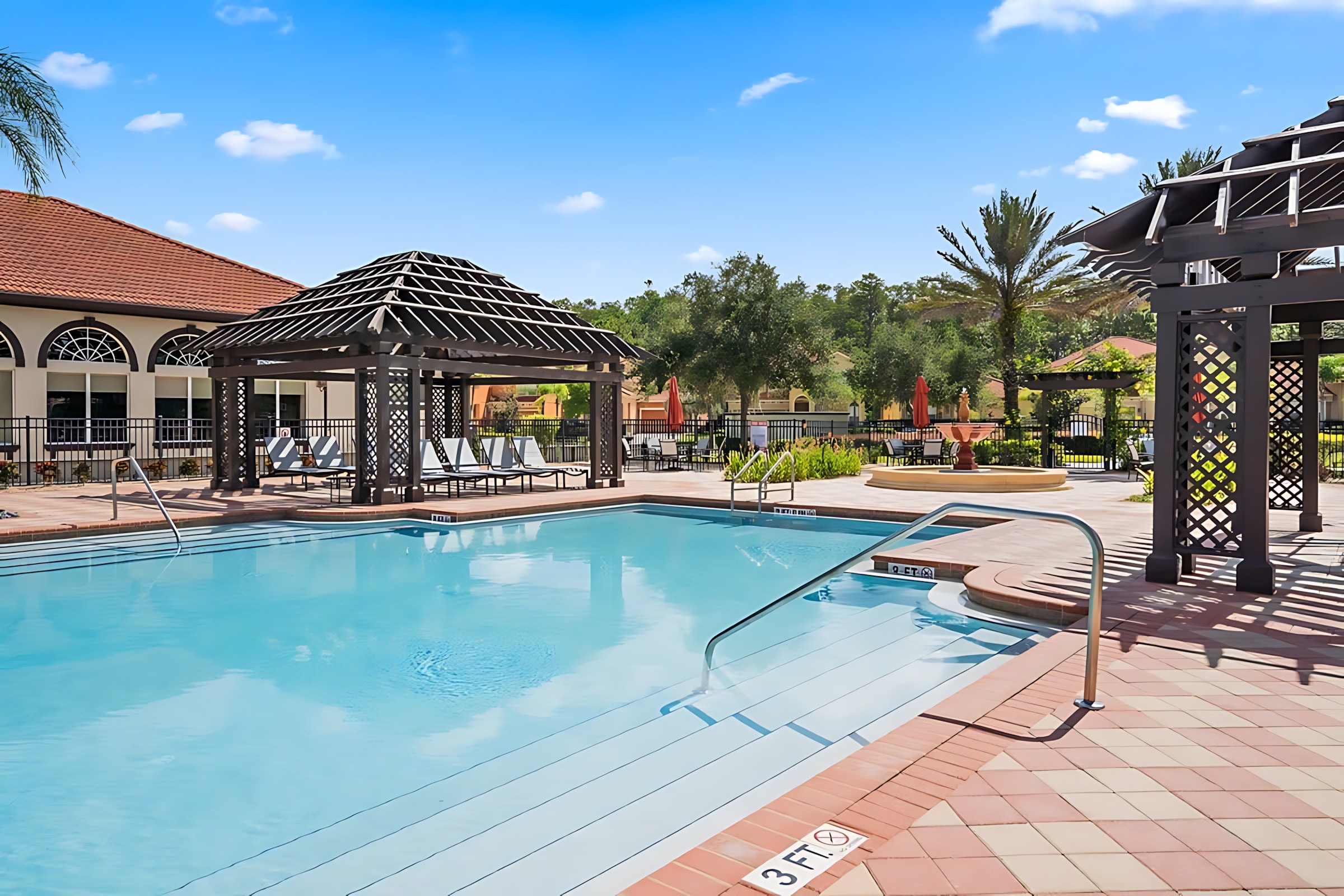 Inviting communal swimming pool with clear blue water, surrounded by a patterned paver deck and shaded pergolas. Steps lead into the pool, and lounge chairs are visible under the pergolas, with a clubhouse in the background.