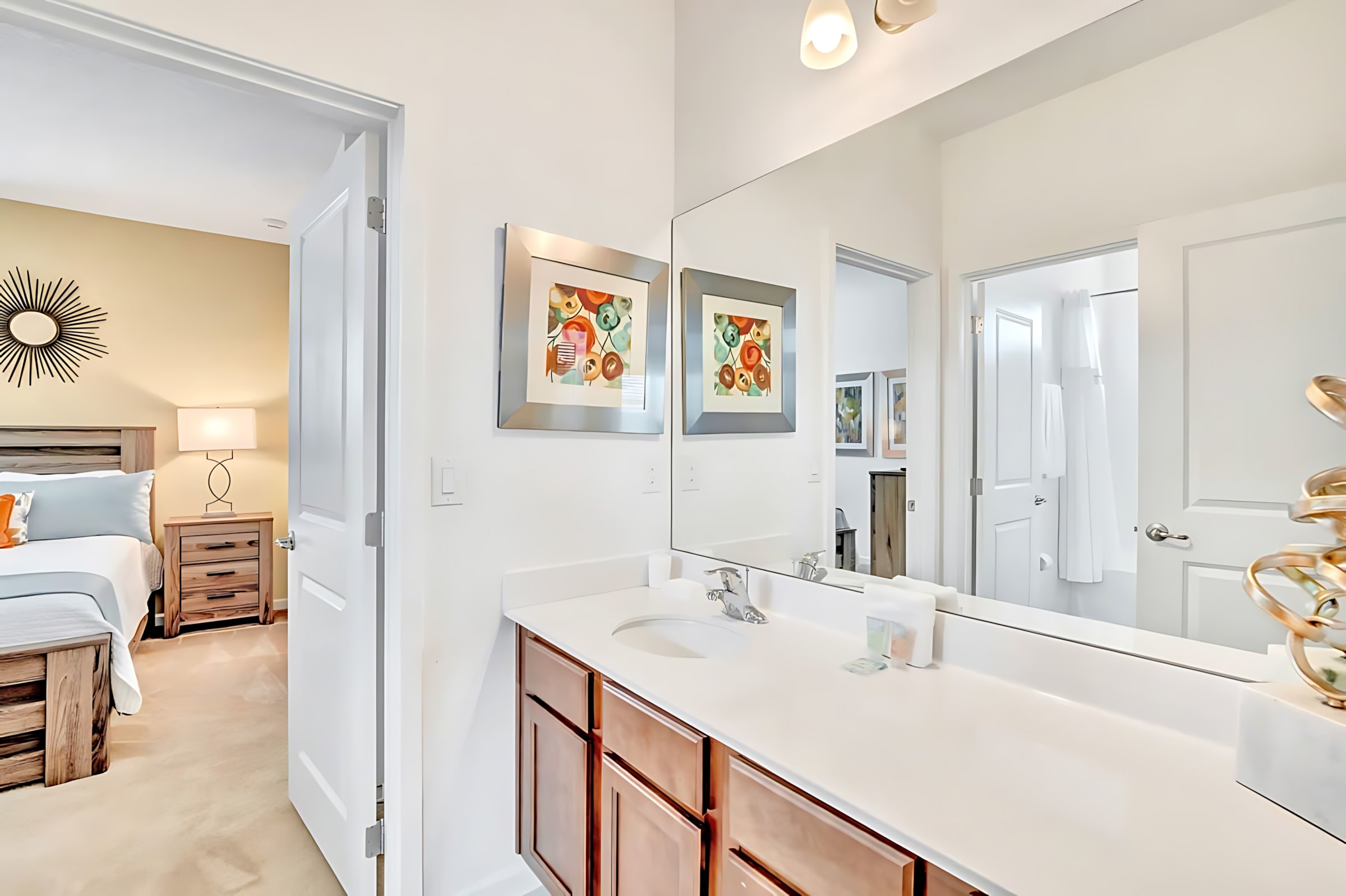 Modern bathroom vanity with a long white countertop, double sinks, and dark wood cabinets below. A large mirror spans the wall above, reflecting the adjacent bedroom and a doorway.