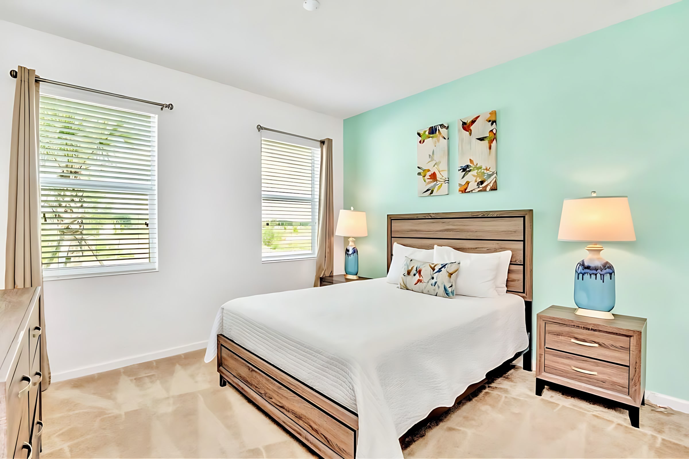 Bright bedroom with a queen bed, wooden headboard, white bedding, and decorative pillows, set against a light mint green accent wall with two bird-themed art pieces. Two windows with blinds are on the white side wall.