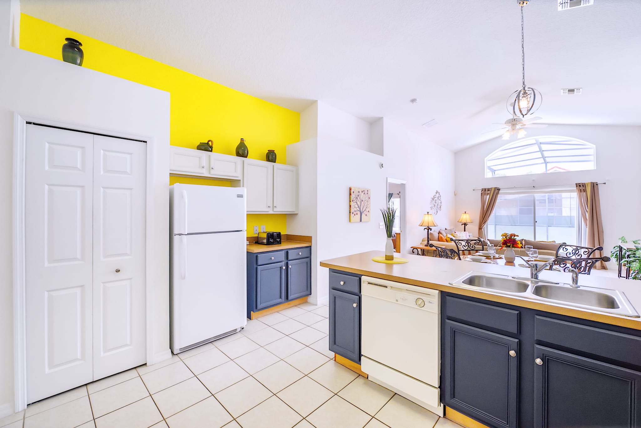 Spacious kitchen with white tiled floors, featuring a white refrigerator, white upper cabinets, dark lower cabinets, and a vibrant yellow accent wall, with an open view towards the living area.
