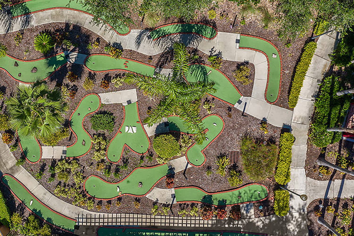 Aerial view of a winding miniature golf course featuring multiple green turf fairways outlined in red, interspersed with concrete pathways and lush tropical landscaping. Various golf holes and obstacles are visible throughout the course.