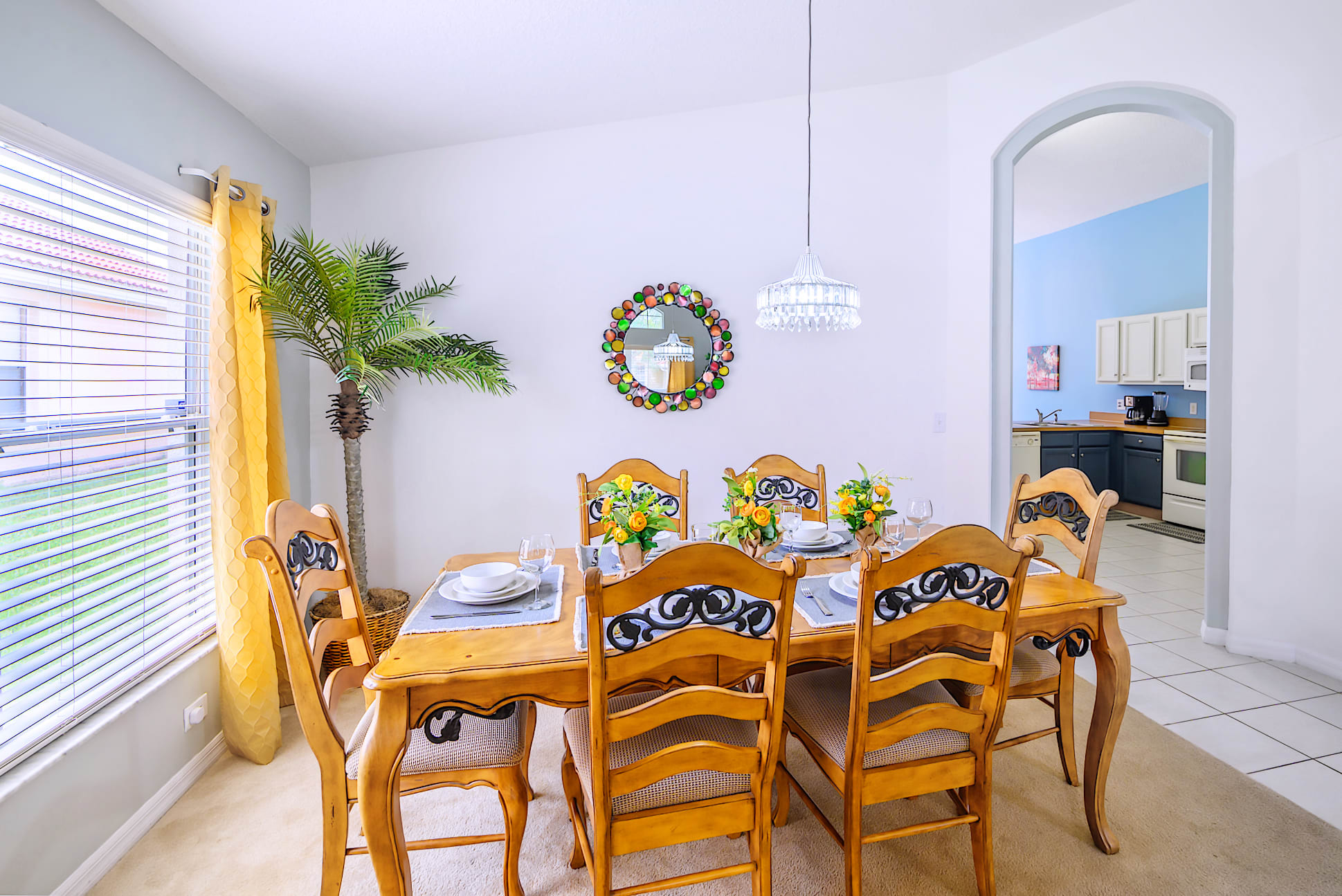Elegant dining area featuring a wooden table set for six with ornate chairs, a decorative round mirror on the wall, and a modern chandelier hanging above, next to a large window with blinds.