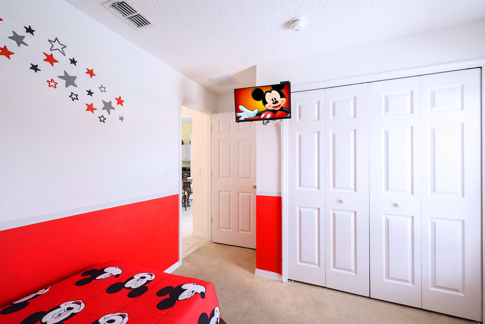 Kids' bedroom with a red and white color scheme, featuring a wall-mounted TV displaying Mickey Mouse, a white bi-fold closet, and a glimpse of a twin bed with Mickey Mouse bedding.