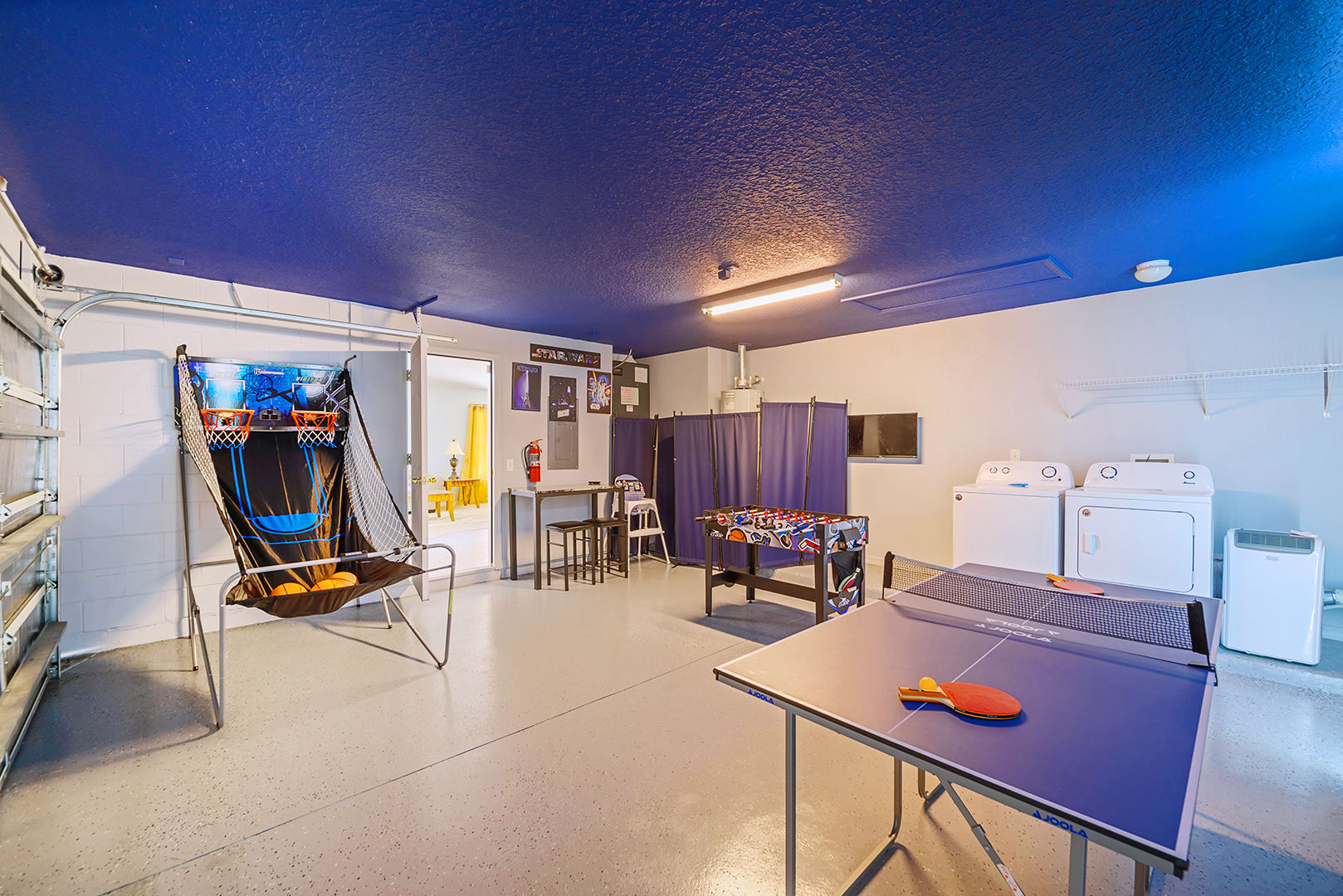 Game room with a blue ceiling, featuring a ping-pong table, a foosball table, a basketball arcade game, and a washer and dryer set against the wall.